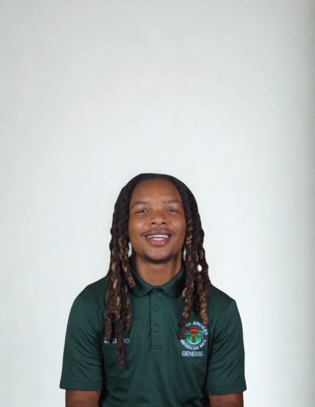 A young man with dreadlocks smiling at the camera, wearing a dark green polo shirt with a logo on the chest against a plain white background.