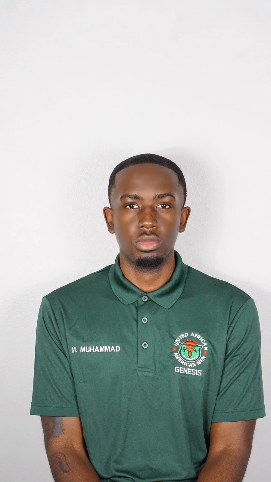 A young man with dark skin and short hair, wearing a green polo shirt with the logo and name of 'United African American Men Genesis' and 'M. Muhammad' embroidered on it, standing against a plain white wall.