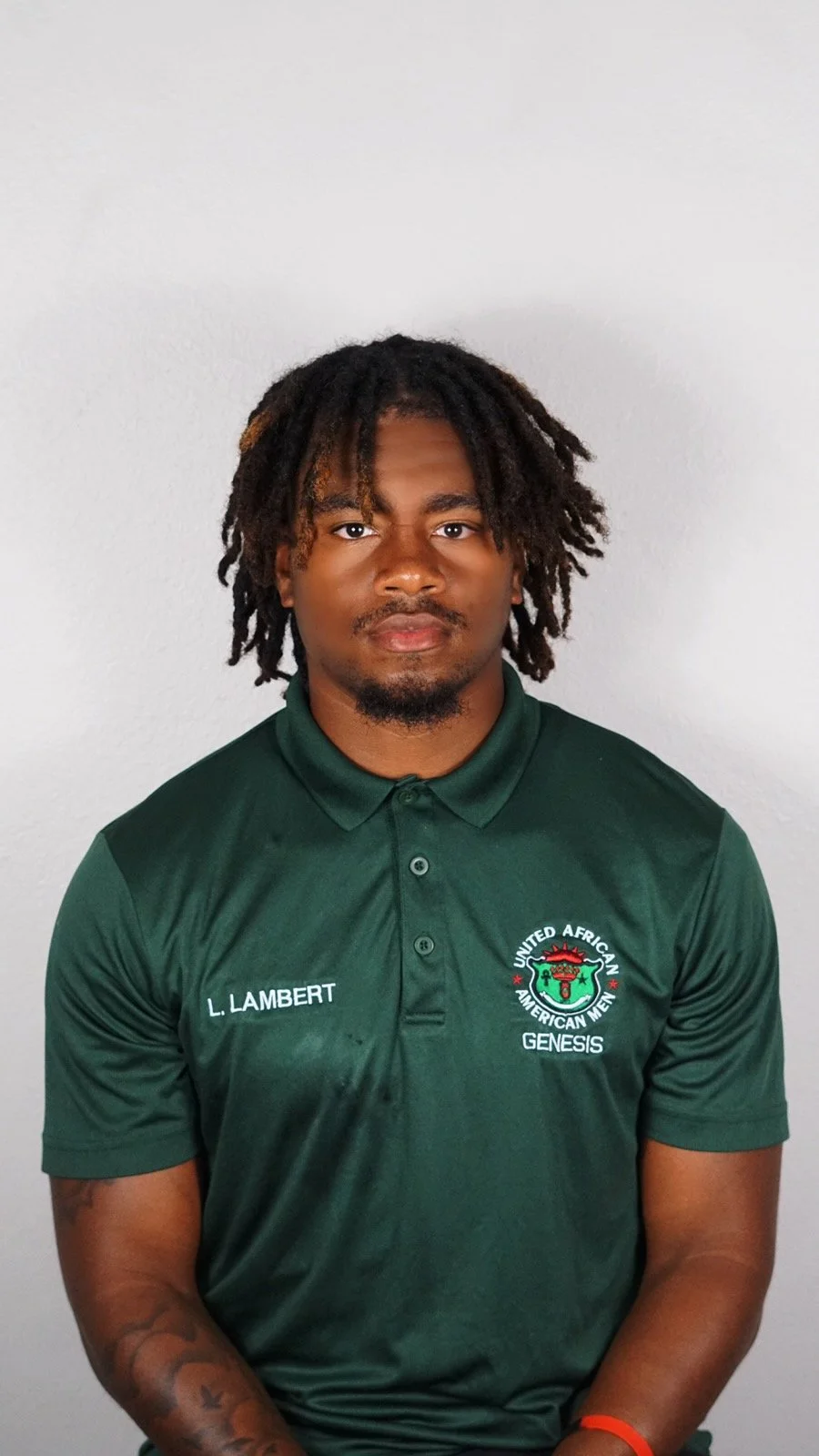 Portrait of a young man with dreadlocks wearing a green polo shirt with a logo and the words 'L. Lambert' and 'United African American Men Genesis'.