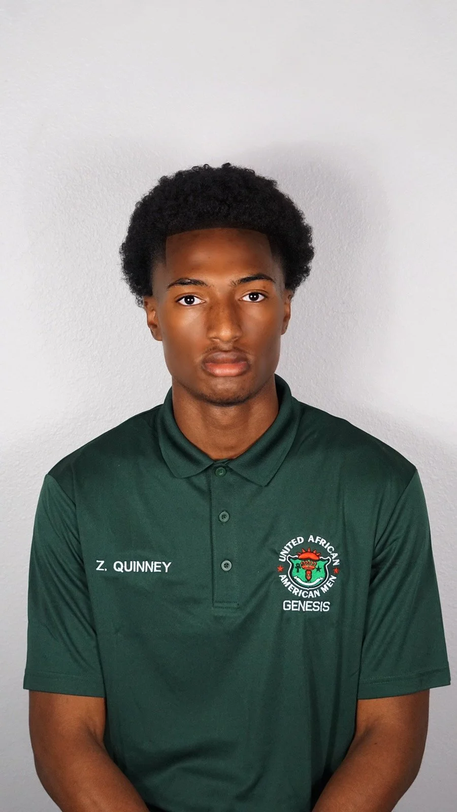 A young man with dark skin and curly black hair wearing a green polo shirt with the logo of United African American Men Genesis and the name Z. Quinney, standing against a plain light gray wall.