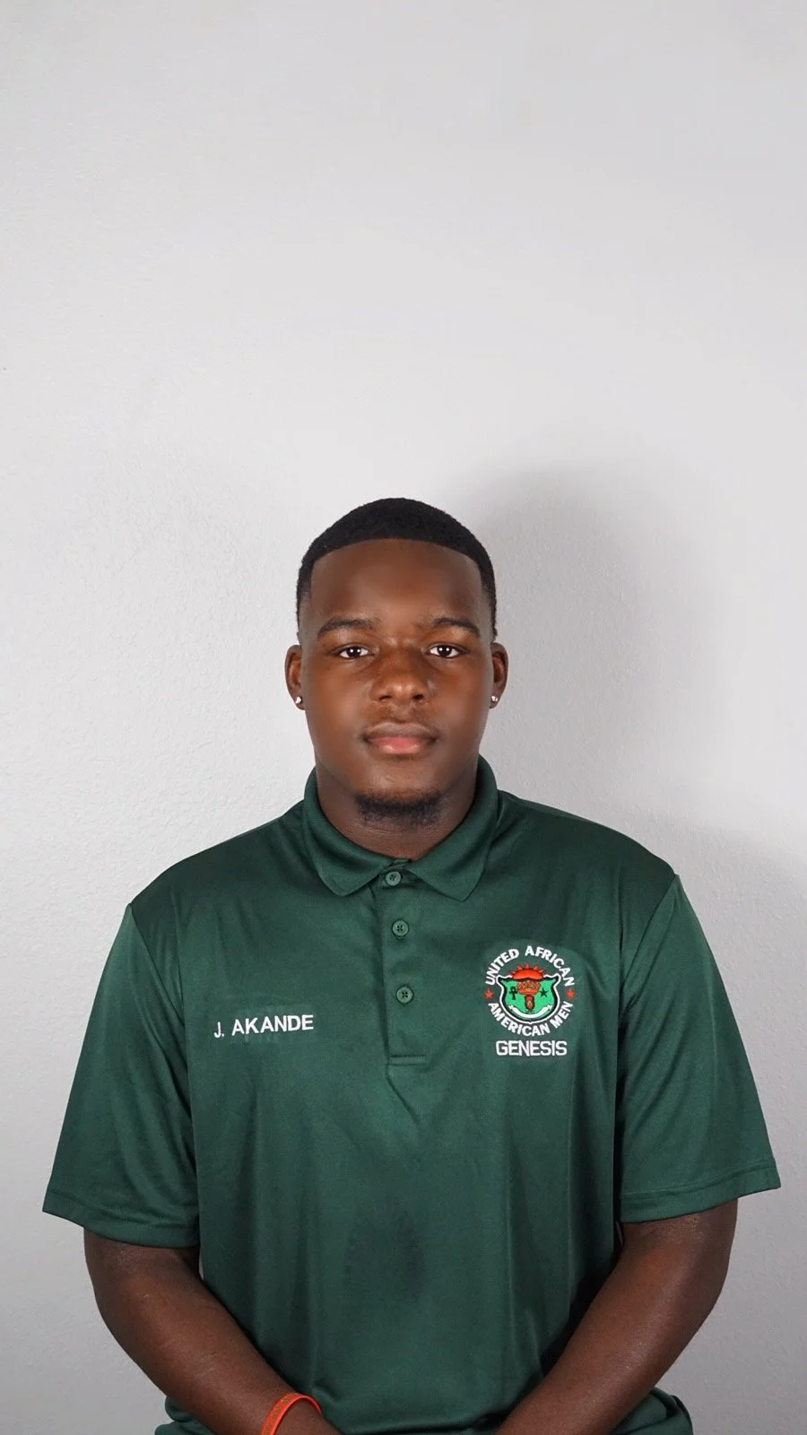 A young man with dark skin and short hair, wearing a green polo shirt with the logo and text 'United African American Men Genesis' and the name 'J. Akande' embroidered on it, standing against a plain light gray wall.