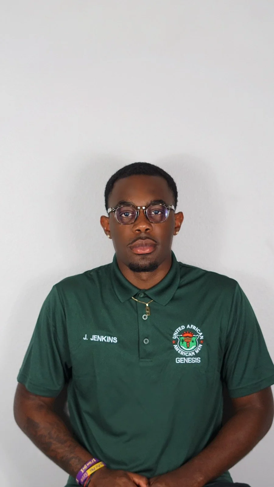 A young Black man wearing glasses and a green polo shirt with logos and text, standing against a plain white wall.