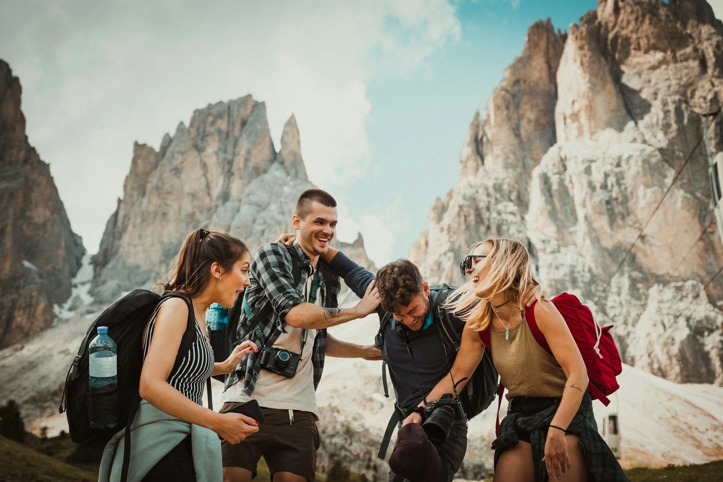 A group of four young friends hiking in the mountains, some with backpacks, cameras, and water bottles, smiling and laughing together in front of rugged mountain peaks.