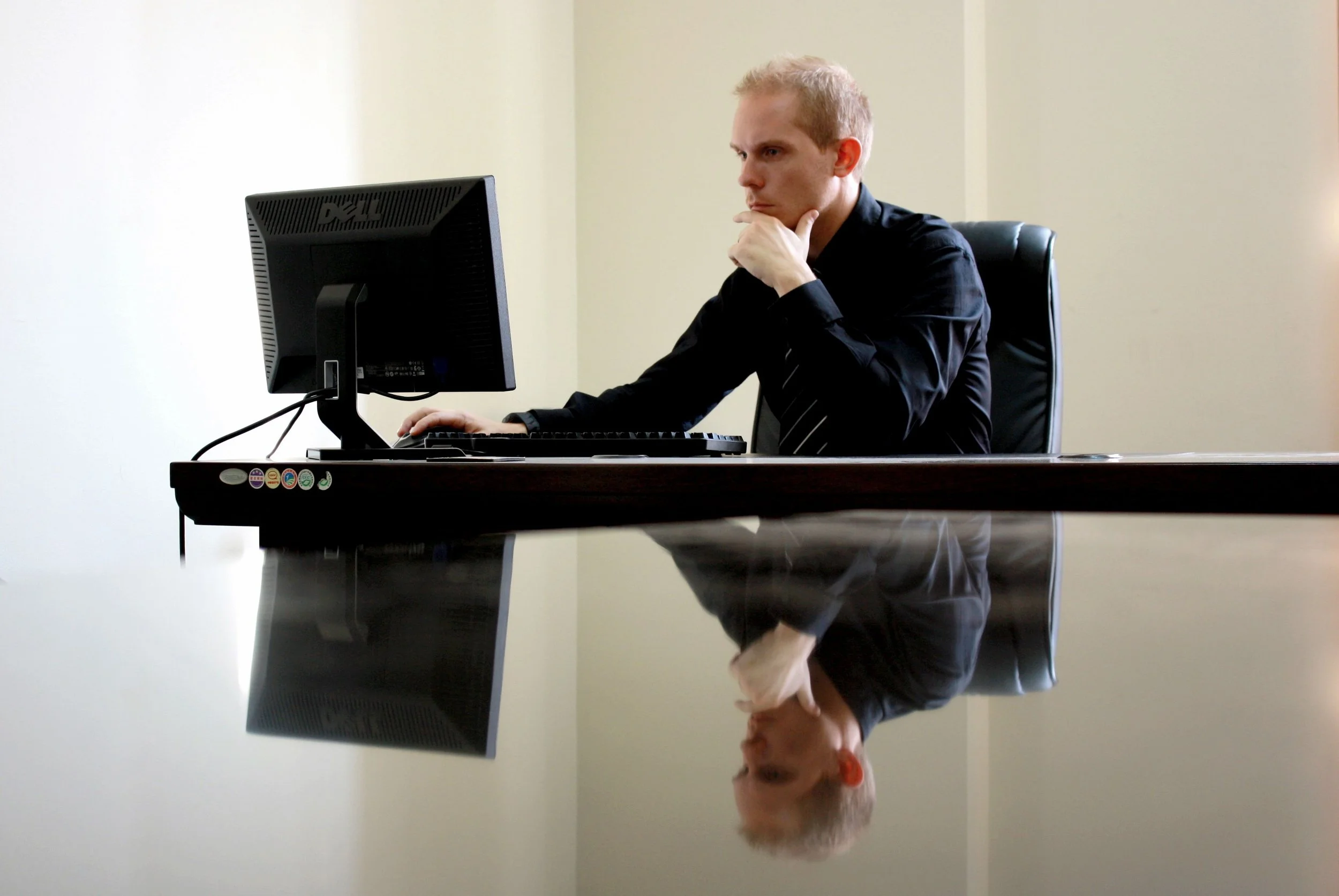 A man in a black shirt and tie sitting at a desk, looking at a computer monitor with his hand on his chin, reflected on a shiny black surface.