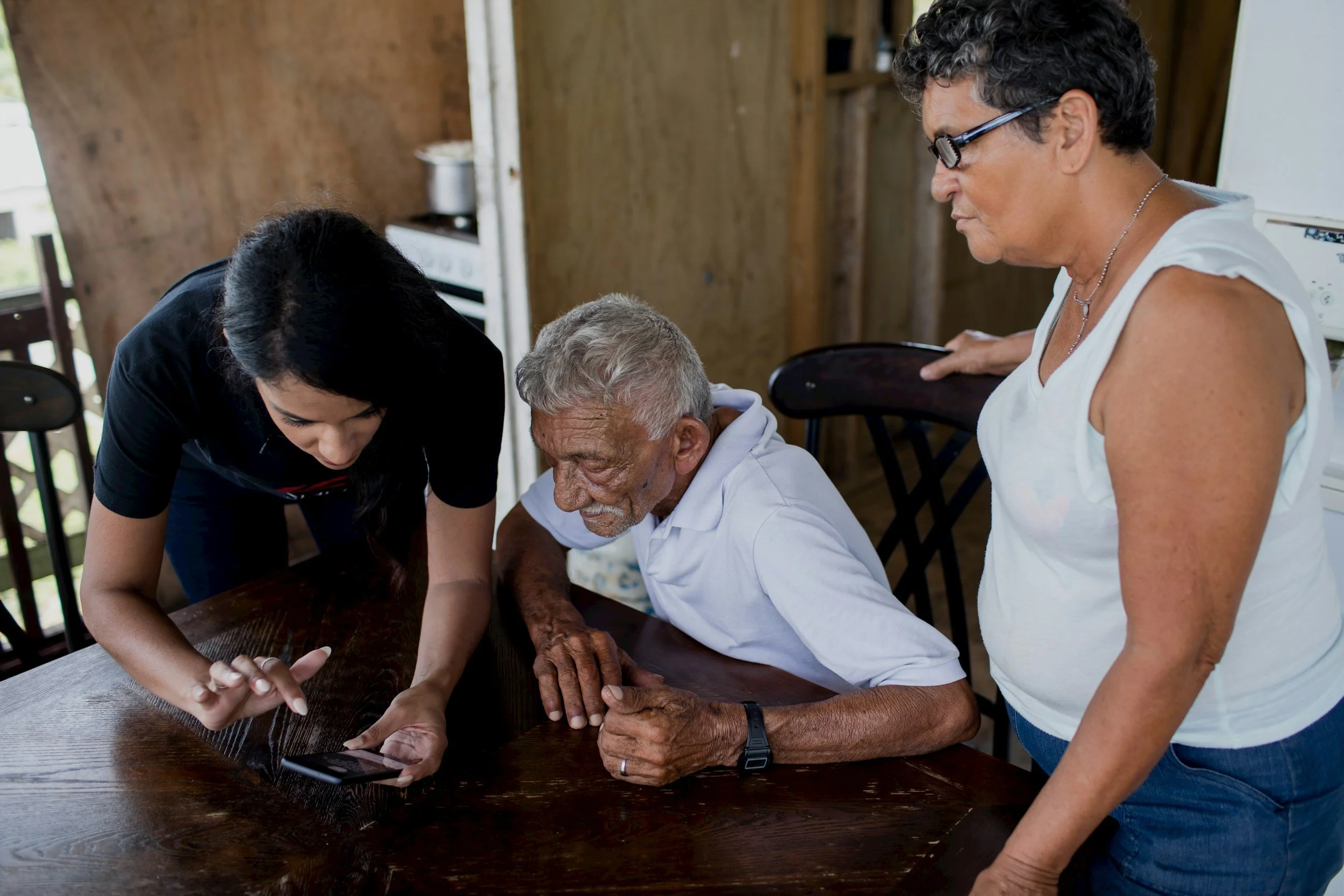 A young woman shows something on a smartphone to an elderly man sitting at a wooden table, with a middle-aged woman standing beside them in a rustic home interior.