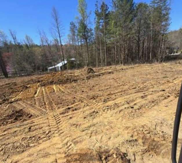 A cleared construction site with tire tracks in the dirt, surrounded by trees and a building in the distance.