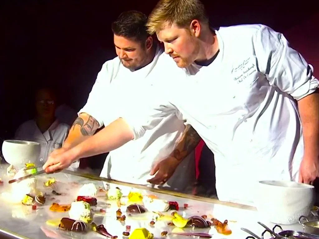 Two chefs in white uniforms are preparing a dessert with chocolate, fruits, and edible decorations on a table, with a few spectators observing in the background.