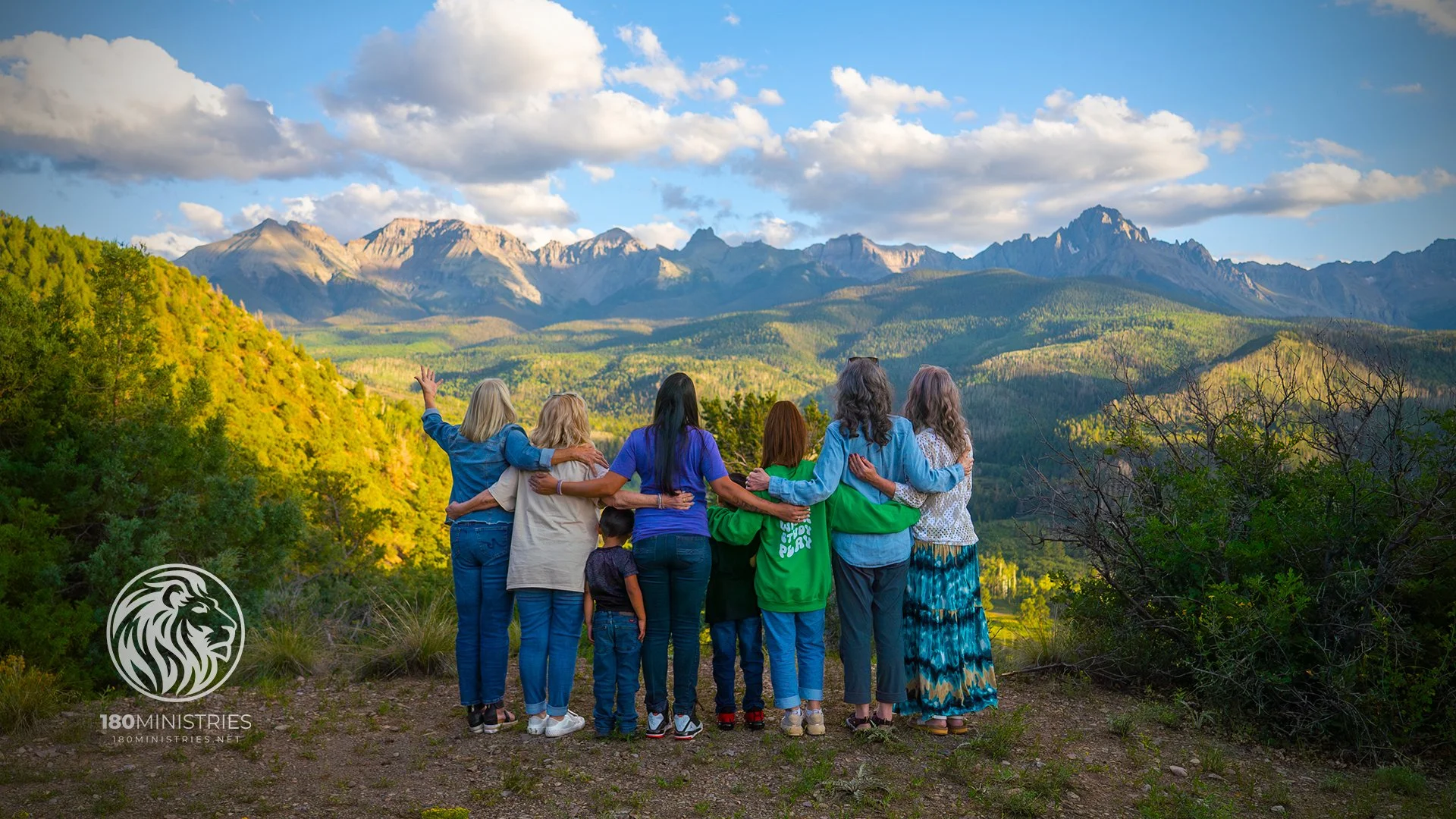Group of diverse people standing outdoors in a scenic mountain landscape, looking at the mountains and clouds, with some embracing each other.