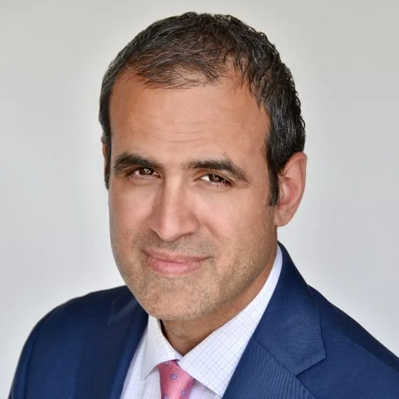 Headshot of a man with short dark hair, wearing a navy blue suit, white shirt, and pink tie against a neutral background.