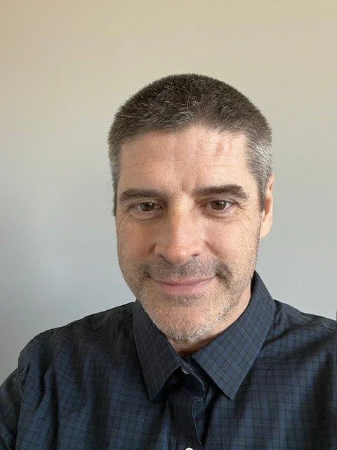 Close-up of a man with short dark hair and light stubble, wearing a dark, checked button-up shirt, smiling slightly, against a plain light-colored background.