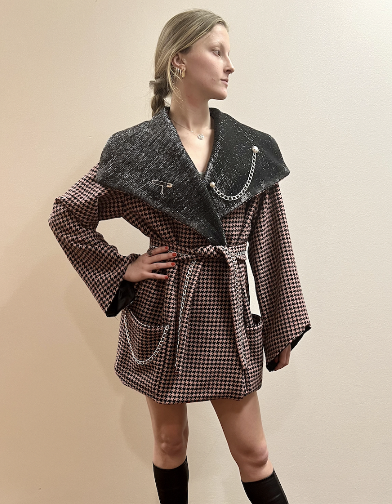 JPA TRENCH – A structured pink houndstooth wool trench with reflective shawl collar detailing and JPA hardware accents. Finished with patch pockets, inverted bell sleeves, a wool belt, and detachable chain draping with JPA charms.