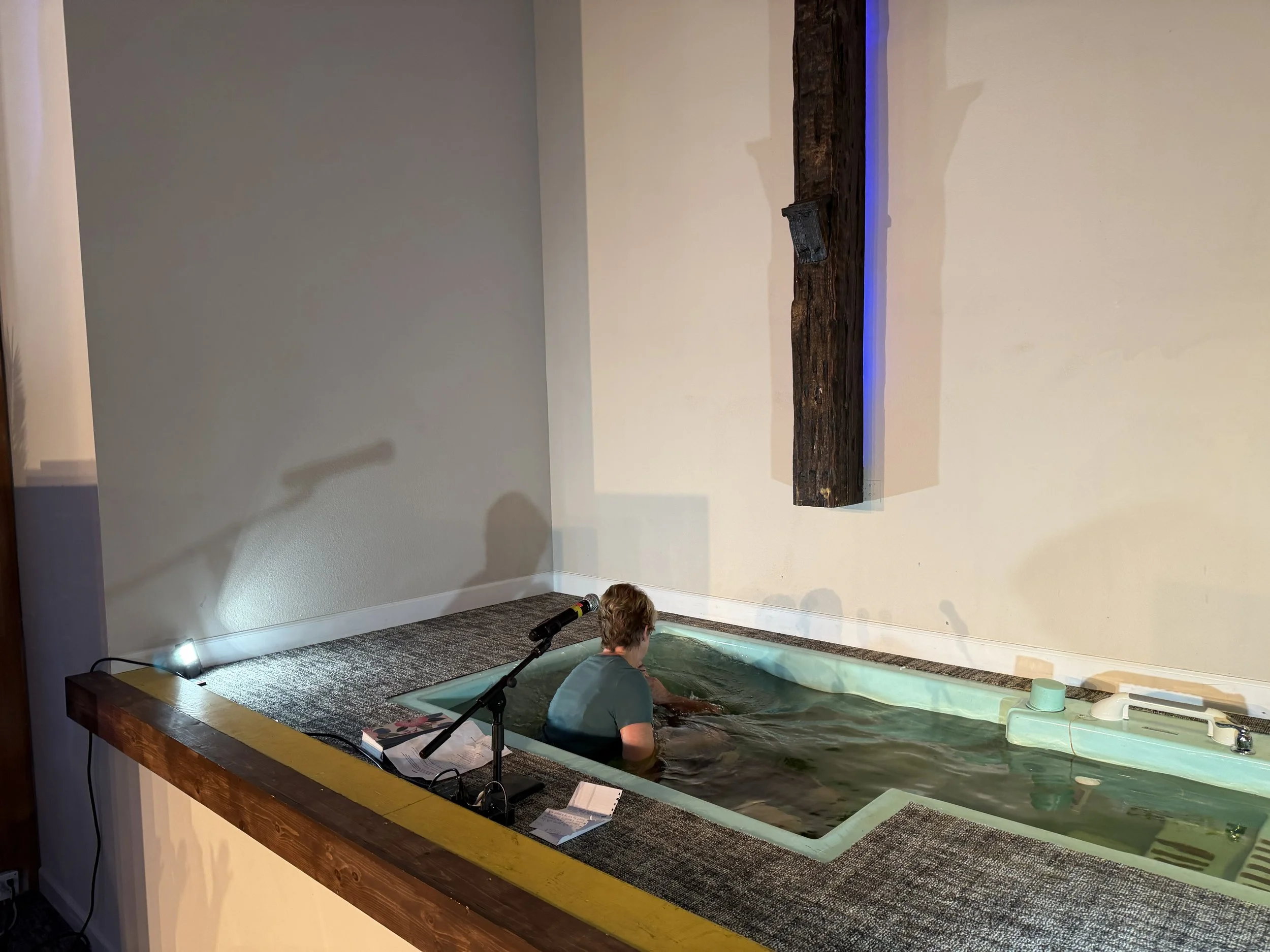 Person sitting in a small, rectangular baptism pool, with a microphone and papers on the edge, in an indoor space with plain walls and a wooden beam.