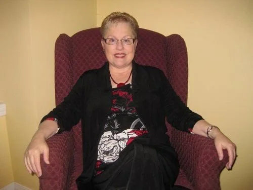 A woman with short blonde hair and glasses sitting in a large, red armchair against a yellow wall.