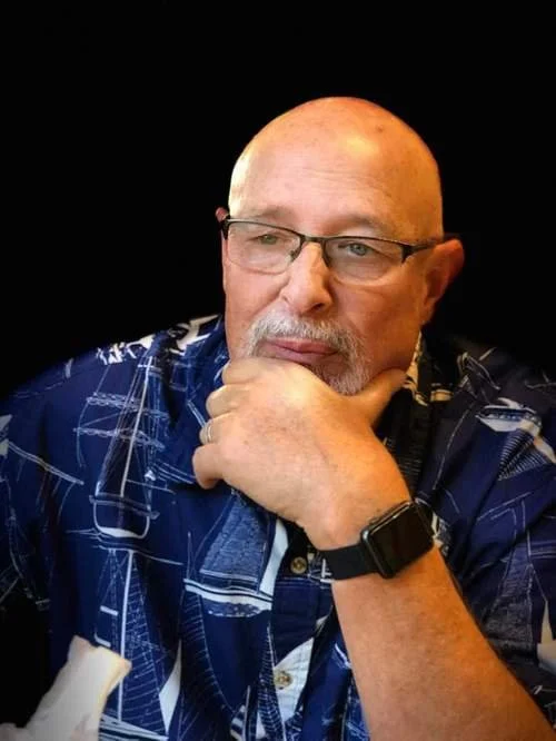 An older man with glasses, a watch, and a gray beard and mustache, wearing a blue patterned shirt, sitting with his hand on his chin against a black background.