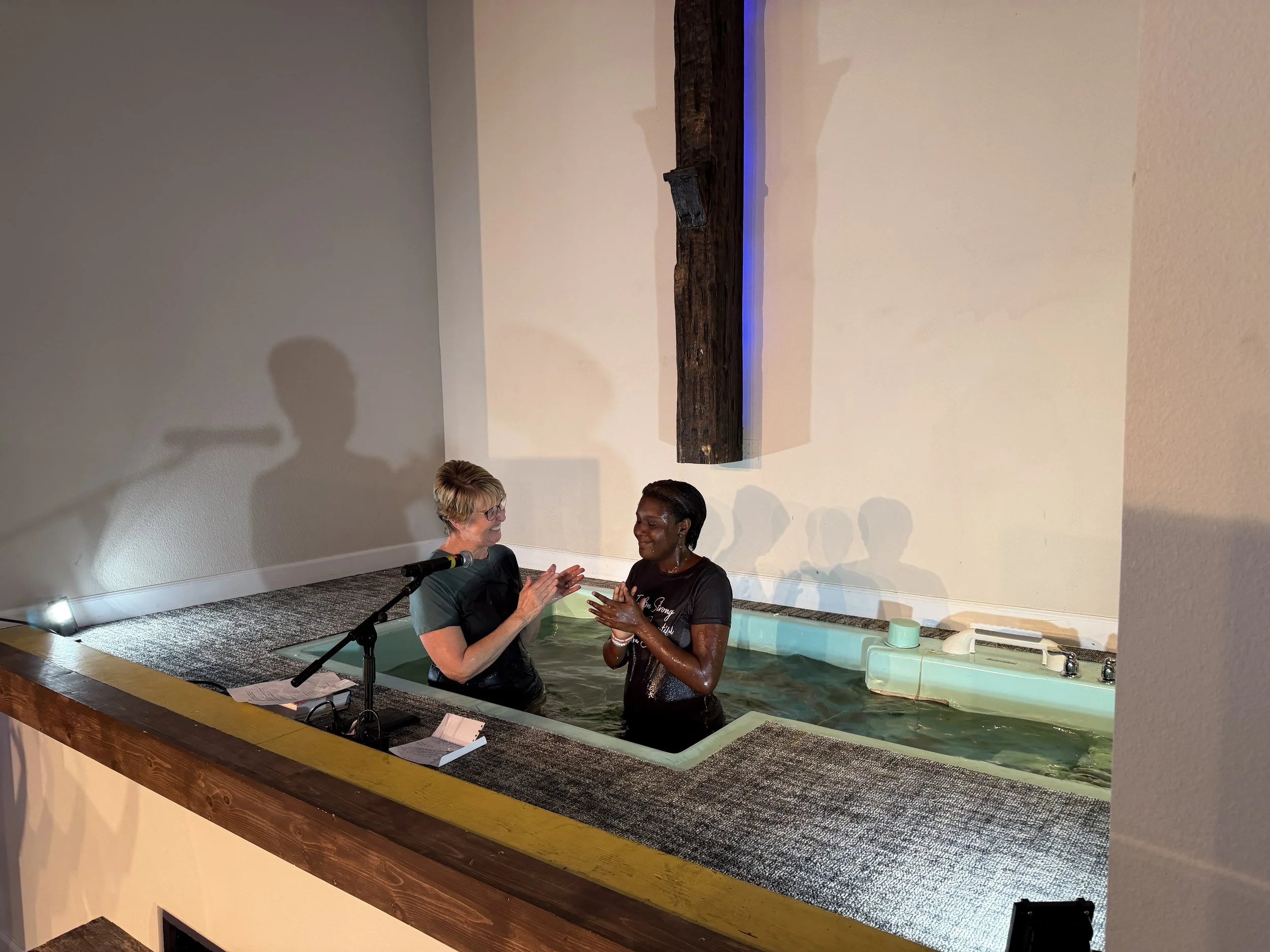 Two women in a baptistry, one clapping, the other with hands together, during a baptism ceremony inside a church.