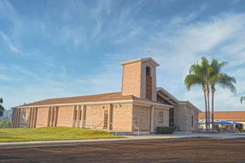 A brick church with a tall tower, surrounded by green grass and palm trees under a partly cloudy sky.