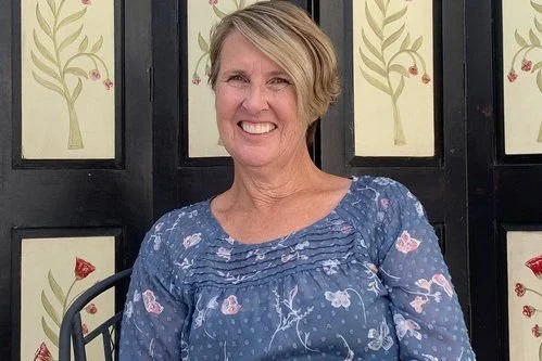 A smiling woman with short blonde hair wearing a blue floral blouse, seated outdoors against a decorative black and cream background.