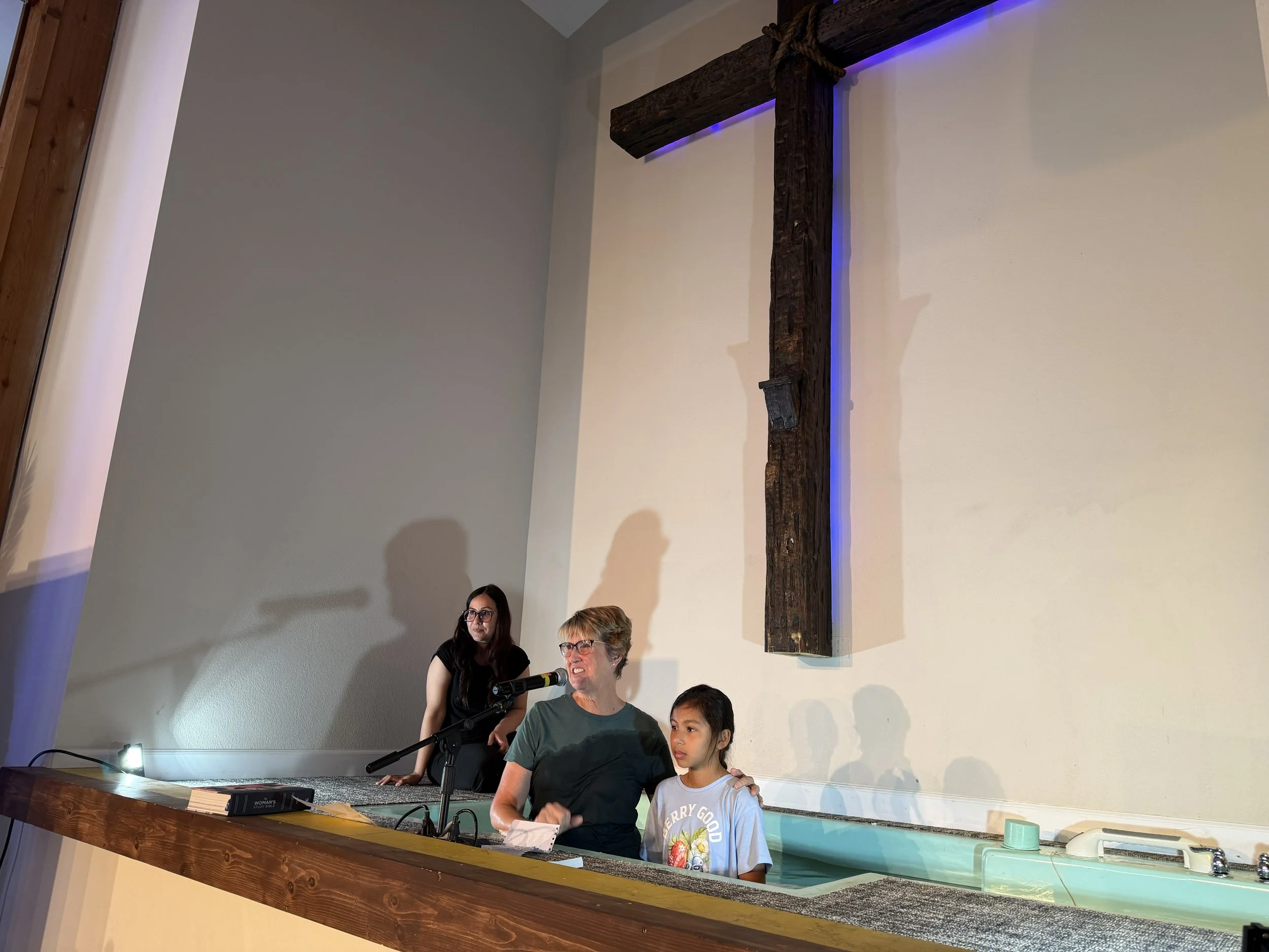 A woman and a young girl stand in front of a church baptismal pool, with a large wooden cross mounted on the wall behind them. The woman is being baptized, and there are two women, one on each side, observing.