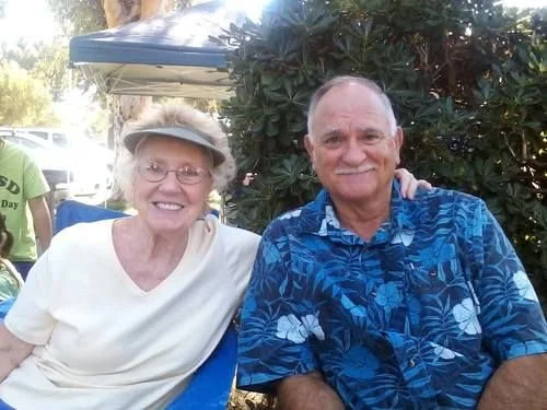 An elderly woman and man sitting outdoors, smiling, with greenery and a blue canopy in the background.