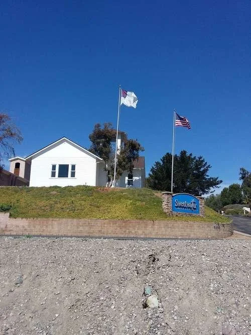 A white house on a small hill with two flags flying, one resembling the Texas flag and the other the American flag. A blue sign with white text reads "Sweetwater." The sky is clear and blue.