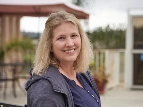 A woman with shoulder-length blonde hair smiling outdoors, wearing a dark jacket, with a blurred background of a porch and trees.