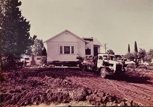 An old black and white photograph of a house with a truck in the foreground on a dirt road.