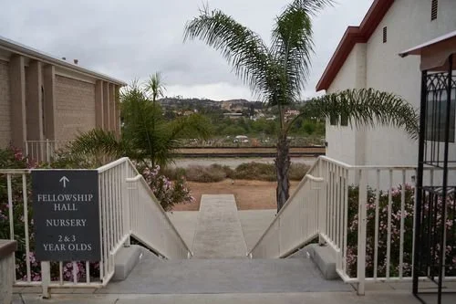 View of a walkway between two buildings with palm trees on either side, leading to an open area with hills in the background. A sign indicates the direction to a nursery for 2-3 year olds in a fellowship hall.