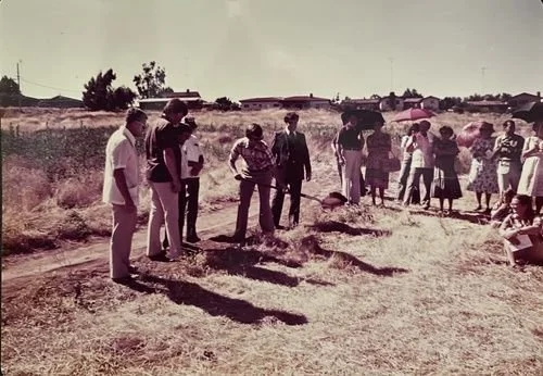 People gathered outdoors, participating in a memorial or solemn event, with some laying flowers on the ground in a rural area.