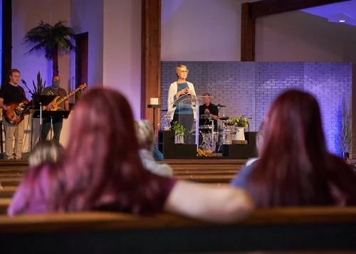 A woman speaking at a church service or event, with a band playing instruments behind her on stage, and an audience watching. The setting appears to be indoors with purple and blue lighting.