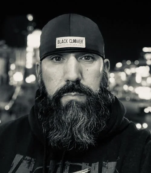 Close-up black and white photo of a man with a beard wearing a bandana with a patch that reads 'Black Clover' on his head, standing outdoors at night with blurred city lights in the background.