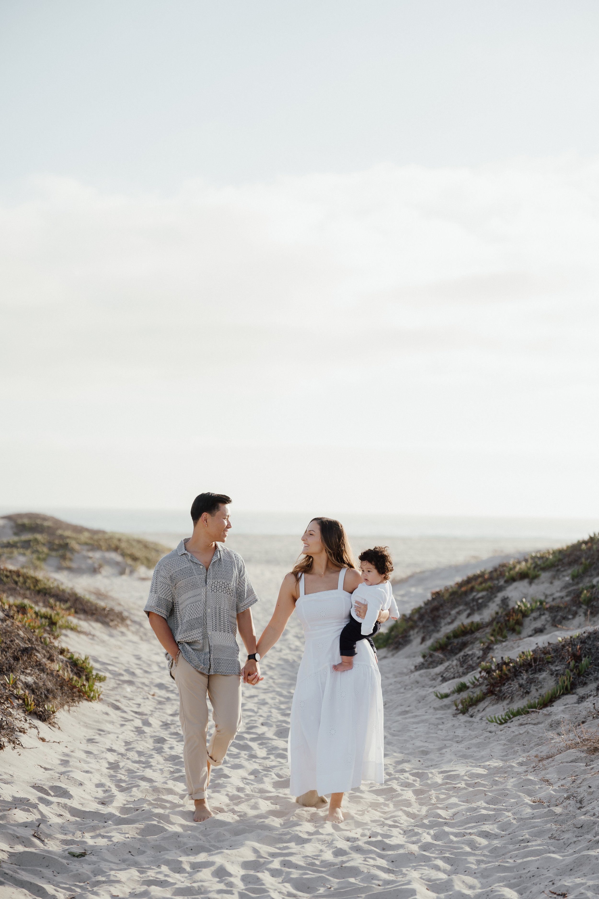 A family of three walking in the dunes at Coronado beach, holding hands and smiling, with sand dunes and a partly cloudy sky in the background.