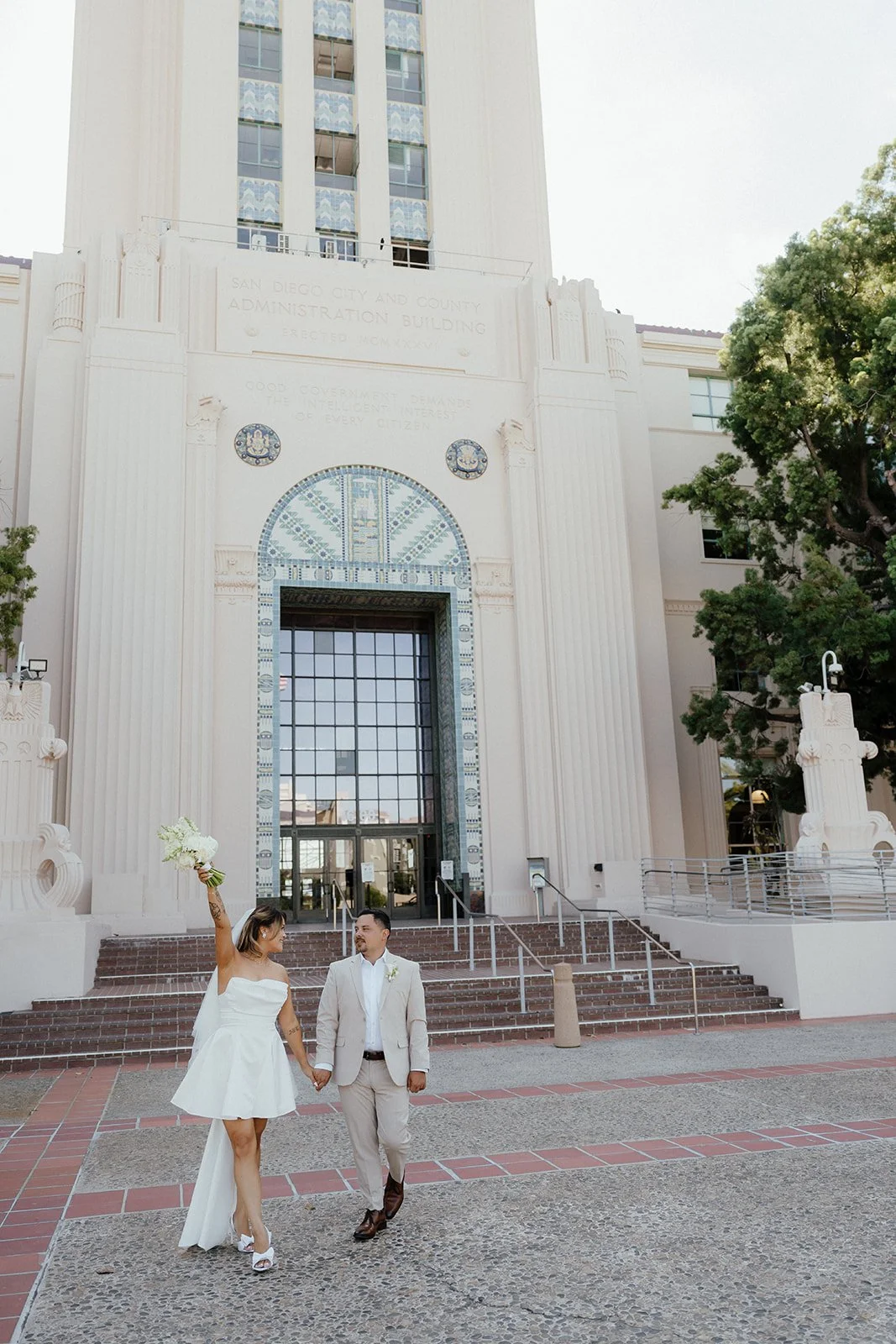 A bride and groom walking hand in hand in front of the San Diego City and County Administration Building. The bride is raising a bouquet of white flowers, and they are both smiling at each other.
