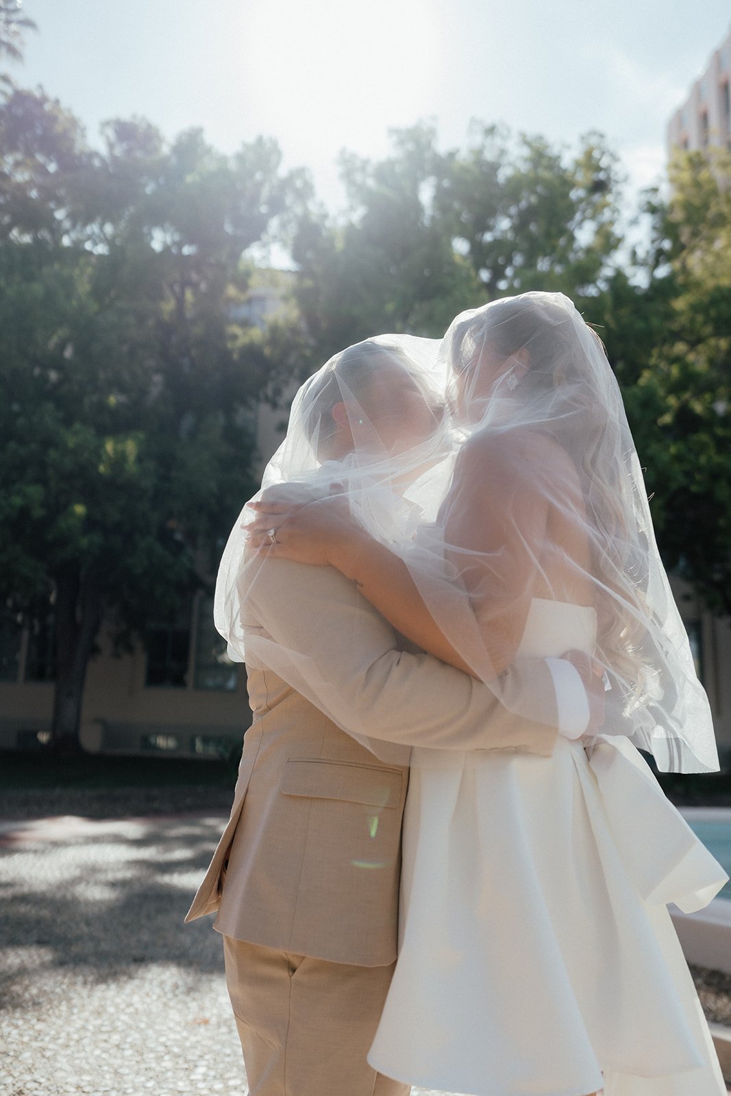 Bride and groom under a bride's veils hugging each other outdoors in sunlight, with trees and buildings in the background during San Diego Elopement.
