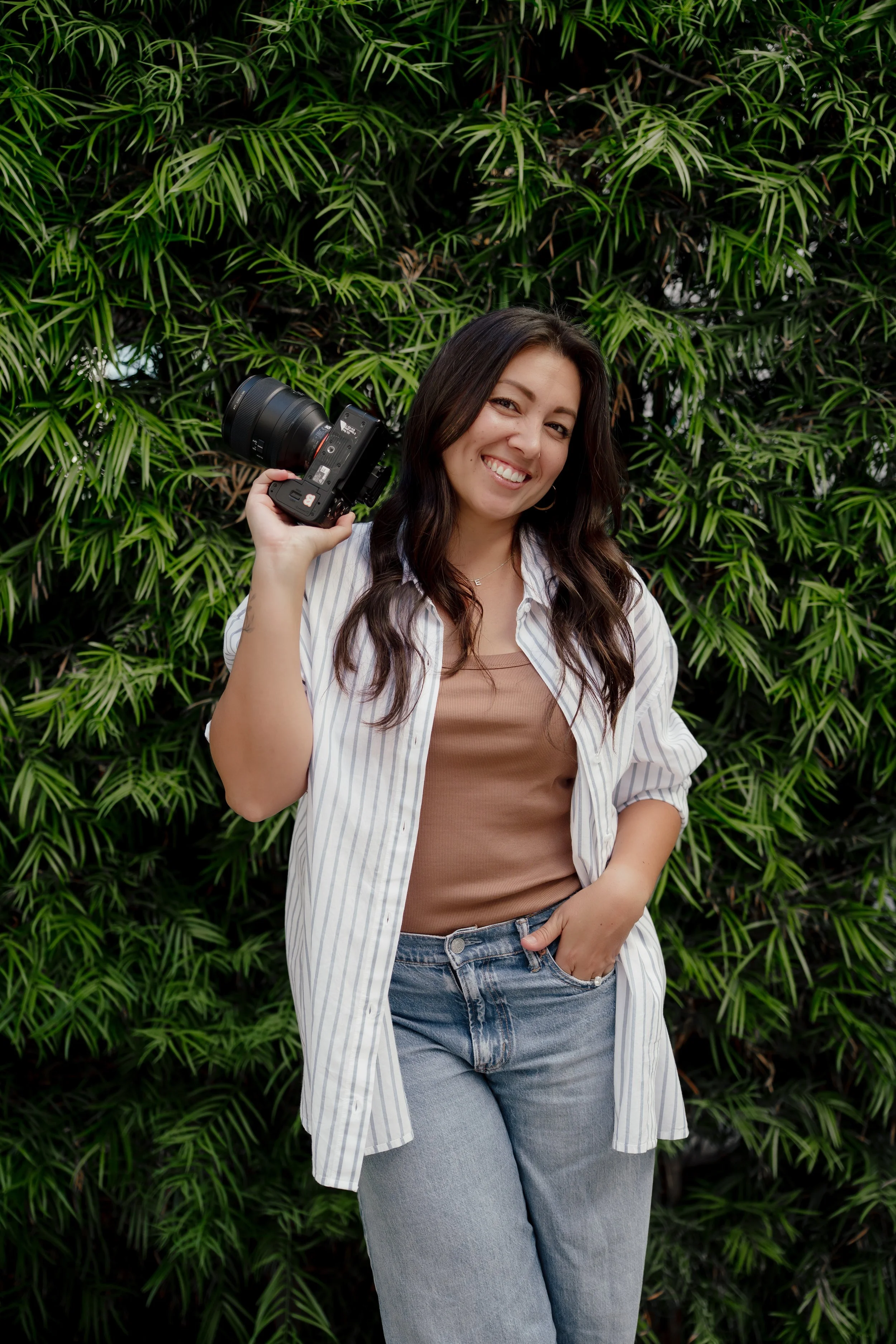 Ally, San Diego wedding and portrait photographer, smiling and holding a camera on her shoulder, standing in front of green foliage.
