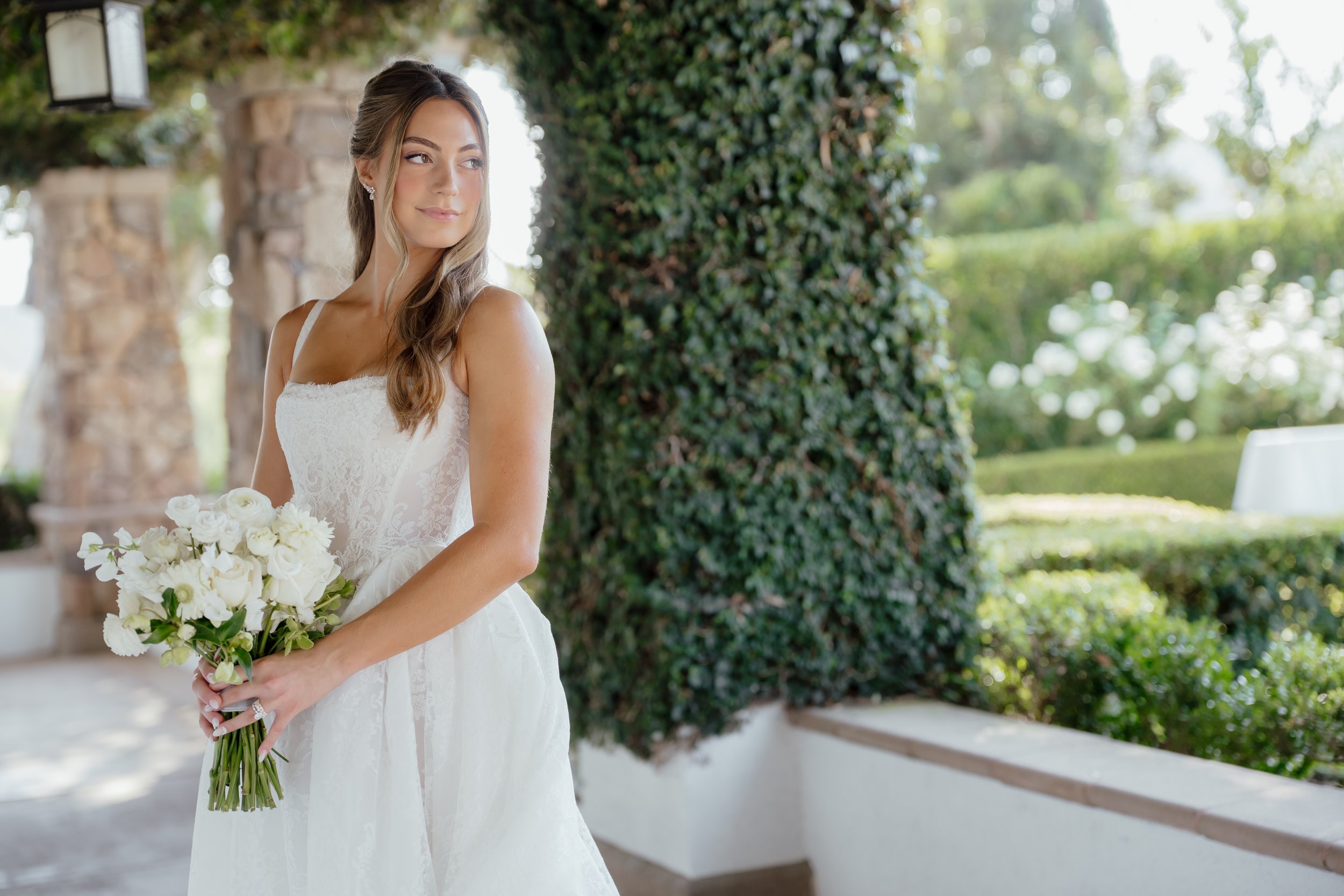 A bride in a white wedding dress holding a bouquet of white flowers, standing outdoors near a stone wall and greenery at Twin Oaks a San Diego wedding venue.