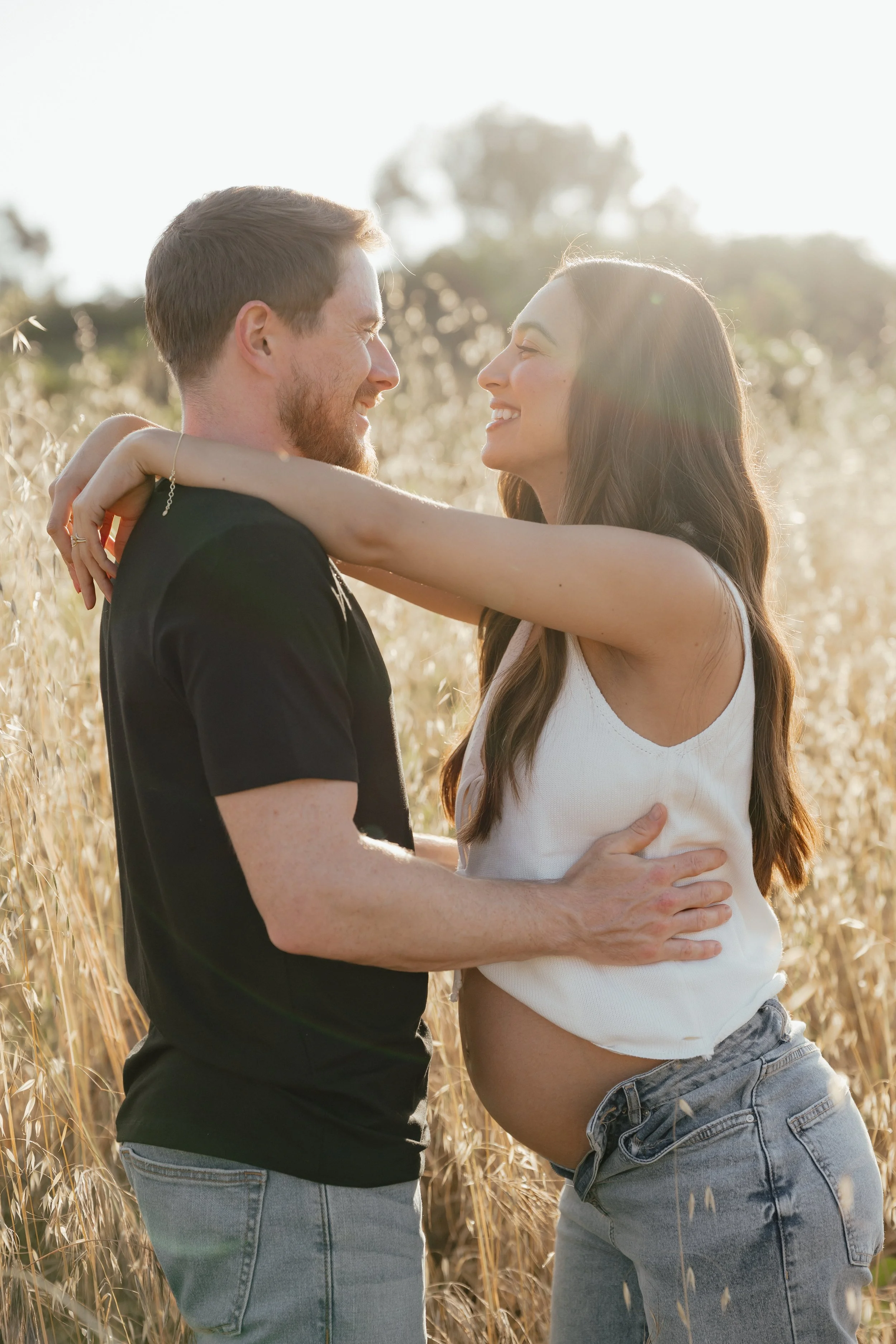 A happy couple embracing in a field of tall, golden grass during sunset, smiling and looking into each other's eyes.