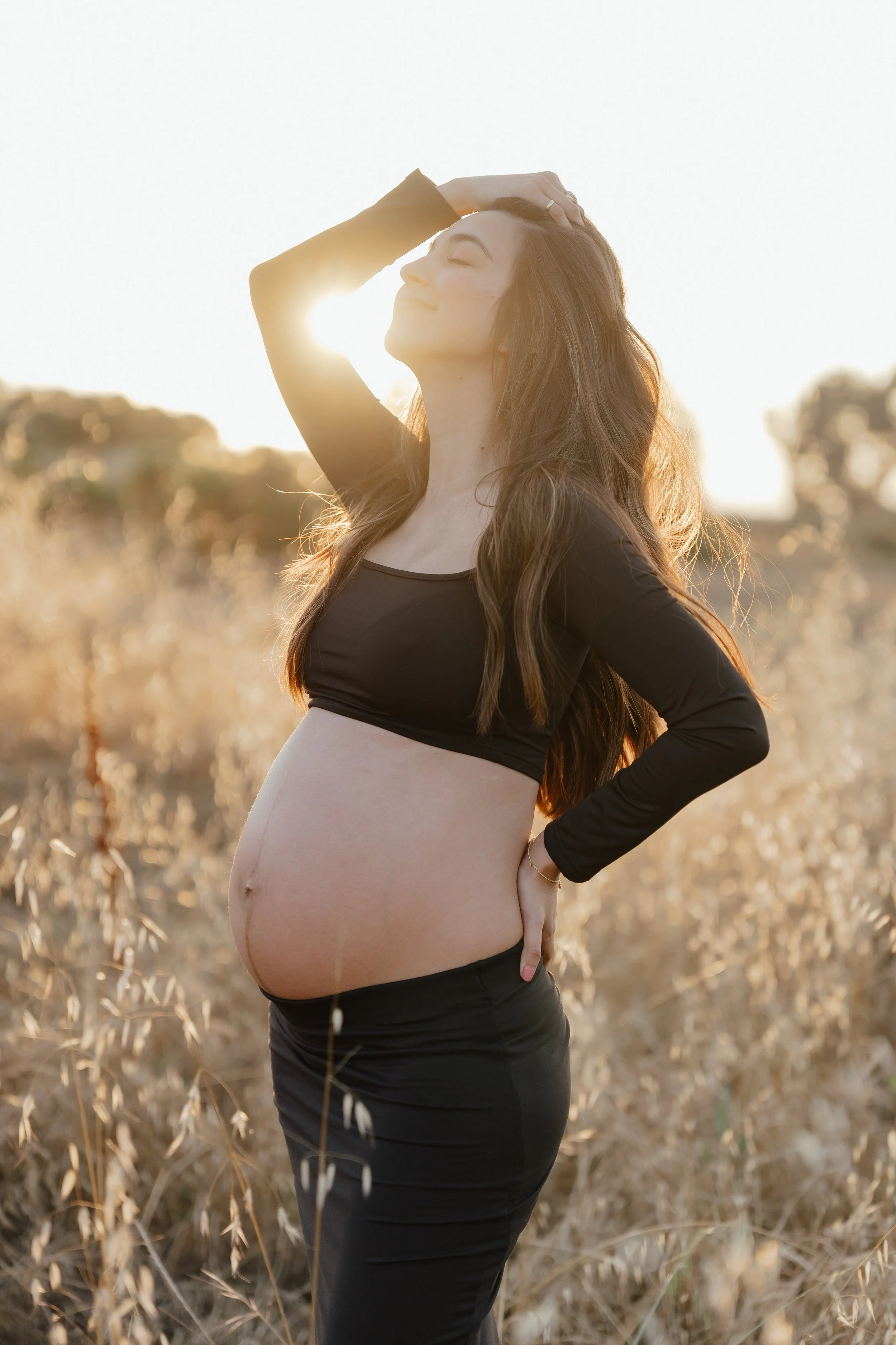 A pregnant woman standing outdoors in a field during sunset, with her eyes closed and one hand on her head, smiling peacefully.