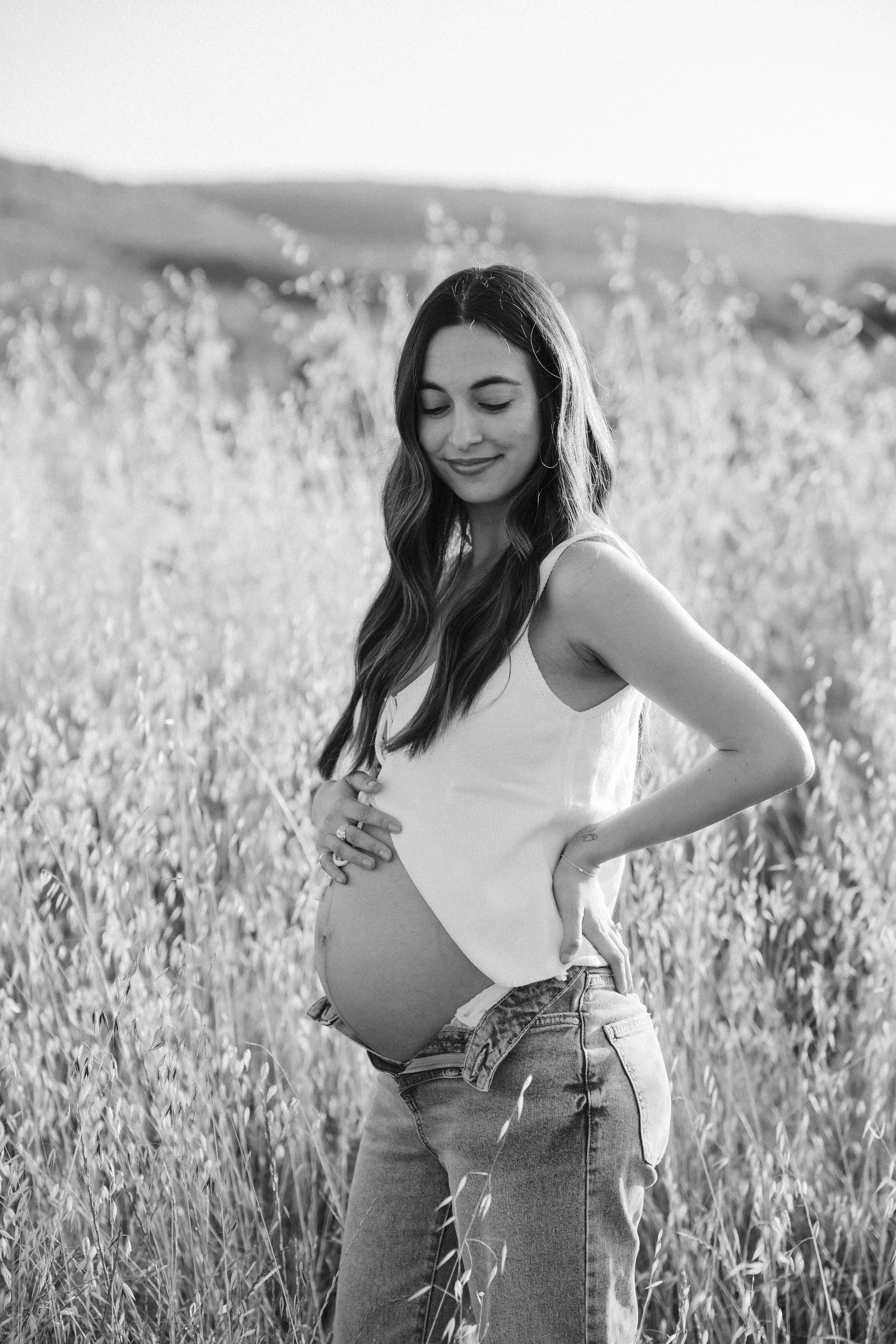 Black and white photo of a pregnant woman standing in a field of tall grass, smiling gently with her eyes closed, holding her belly with one hand and resting her other hand on her hip.
