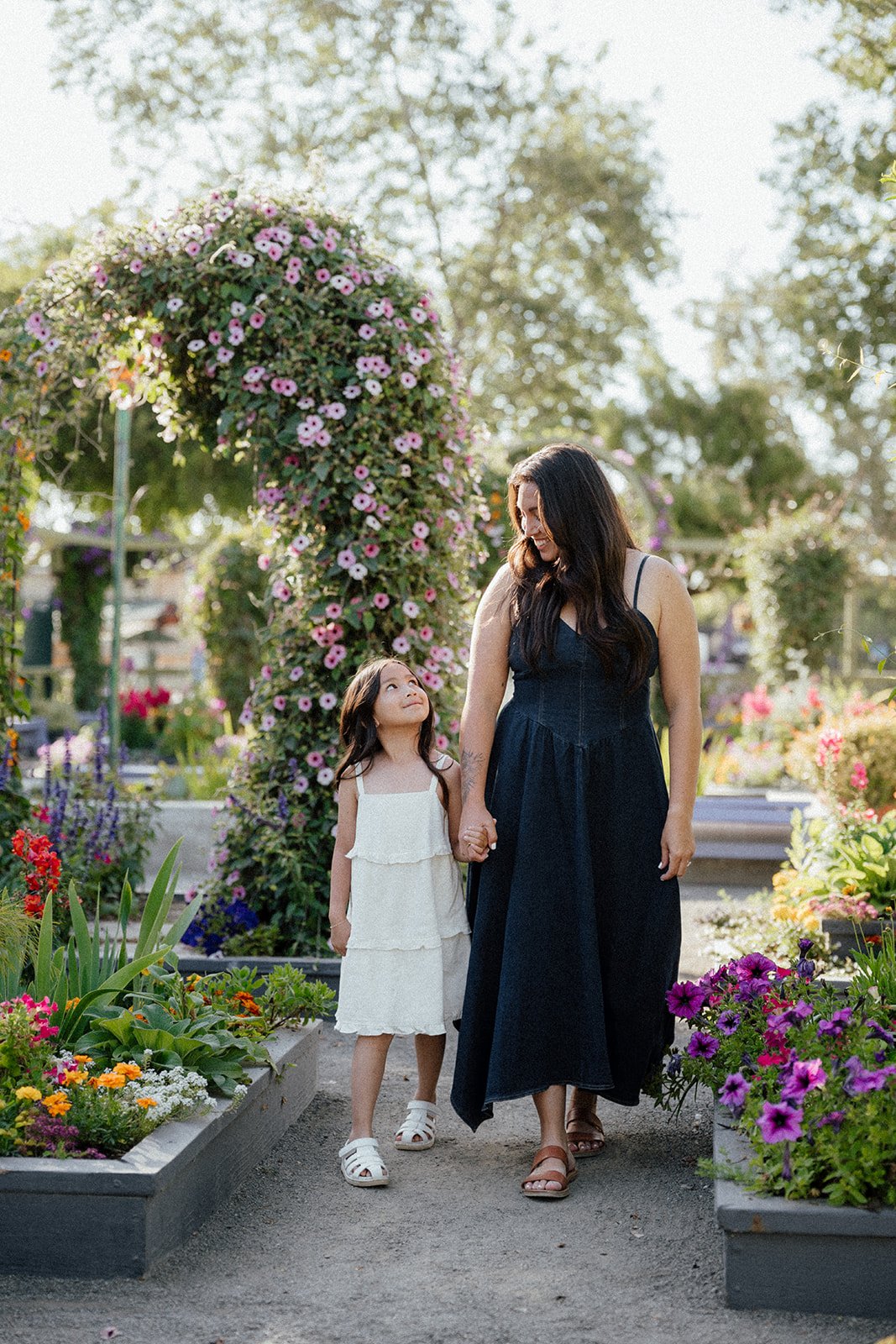 A woman and a young girl hold hands and walk through a flower garden. The woman wears a dark dress and the girl wears a white dress. They look at each other smiling, surrounded by colorful flowers and pink flowering vines.