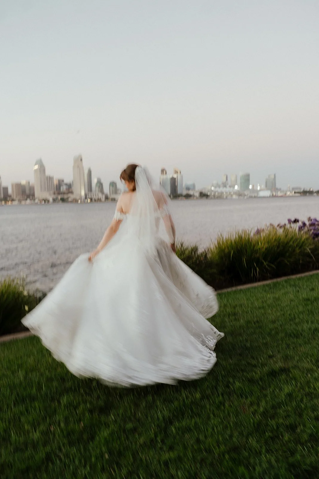 Bride in a white wedding gown and veil turning by a river with a city skyline in the background during sunset during San Diego Elopement.