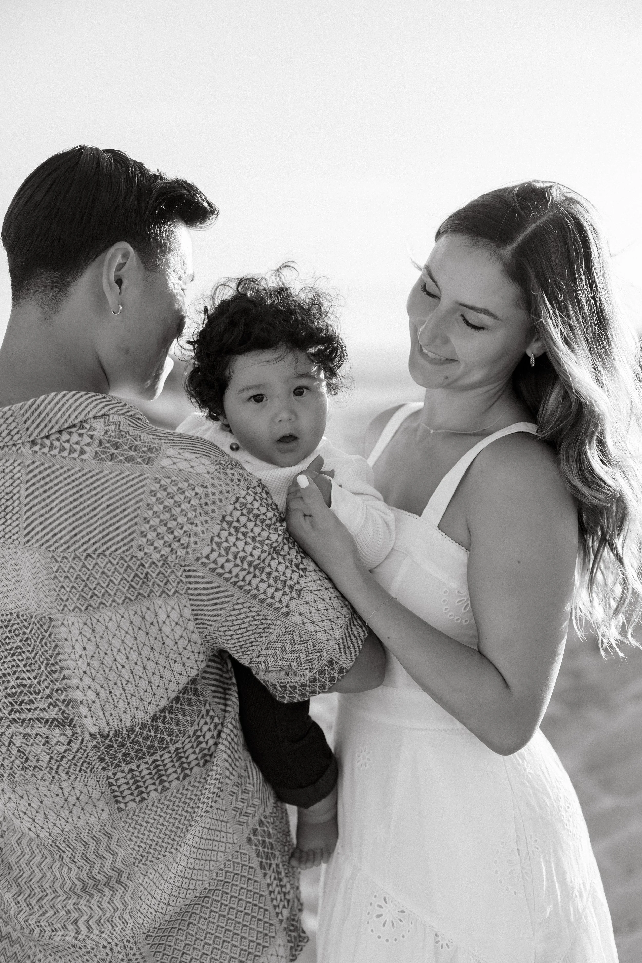 A happy woman holding a young child with curly hair, while a man looks at them, outdoors in a natural setting with bright sunlight.