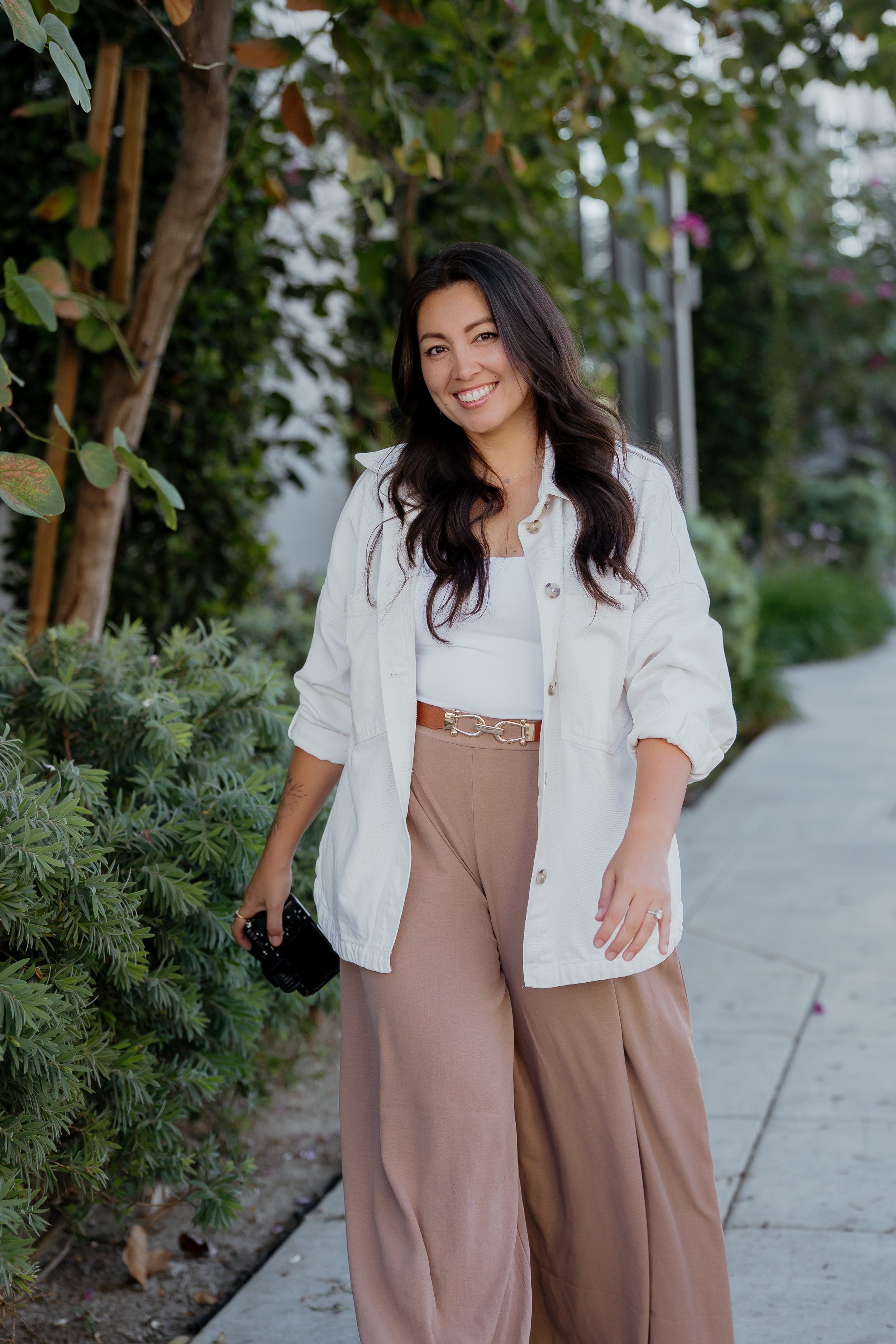Ally, owner of Seagrave Photography, smiling while walking outdoors on a sidewalk, surrounded by lush green plants and trees, holding a camera in her right hand.