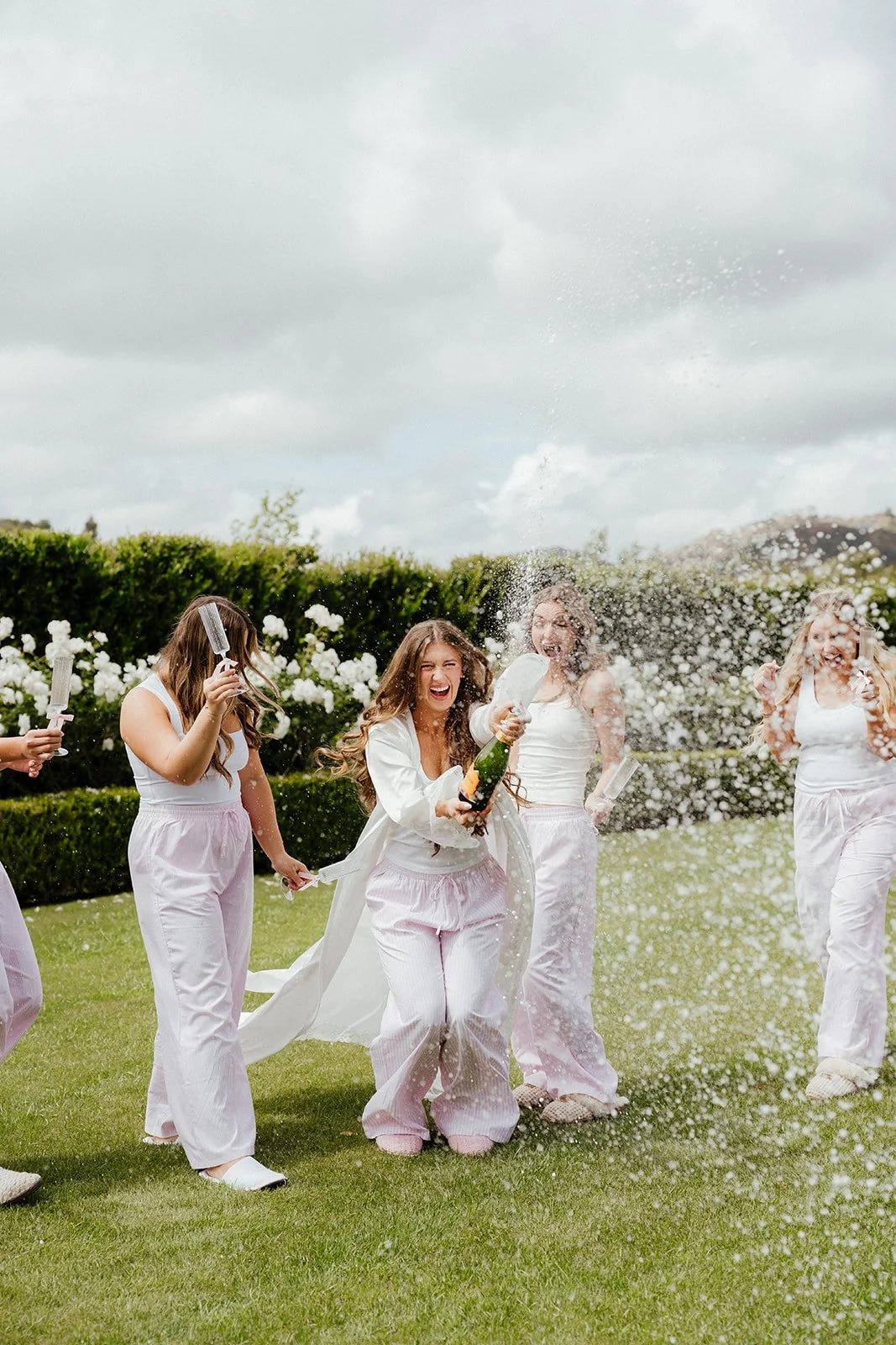 Group of women celebrating outdoors with champagne, wearing white pajamas, laughing, and spraying champagne at Twin Oaks Golf Course Wedding Venue.