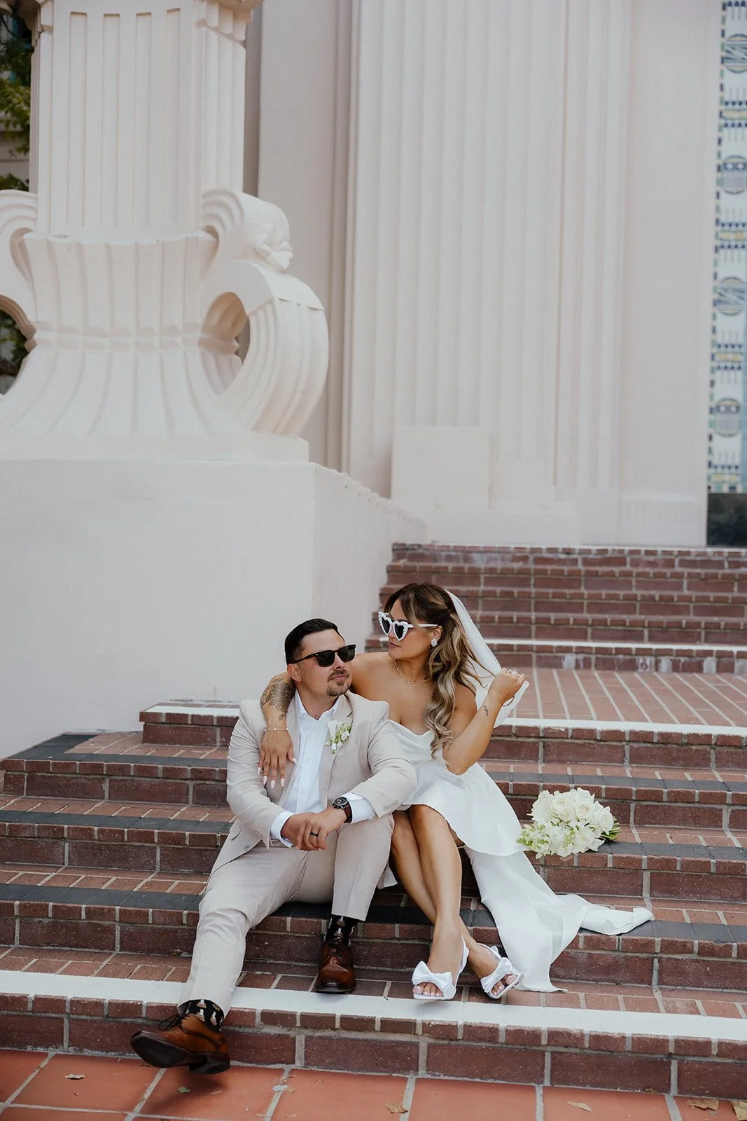 A bride and groom sitting on brick steps outside a building with white columns and architecture at their Downtown San Diego Elopement.
