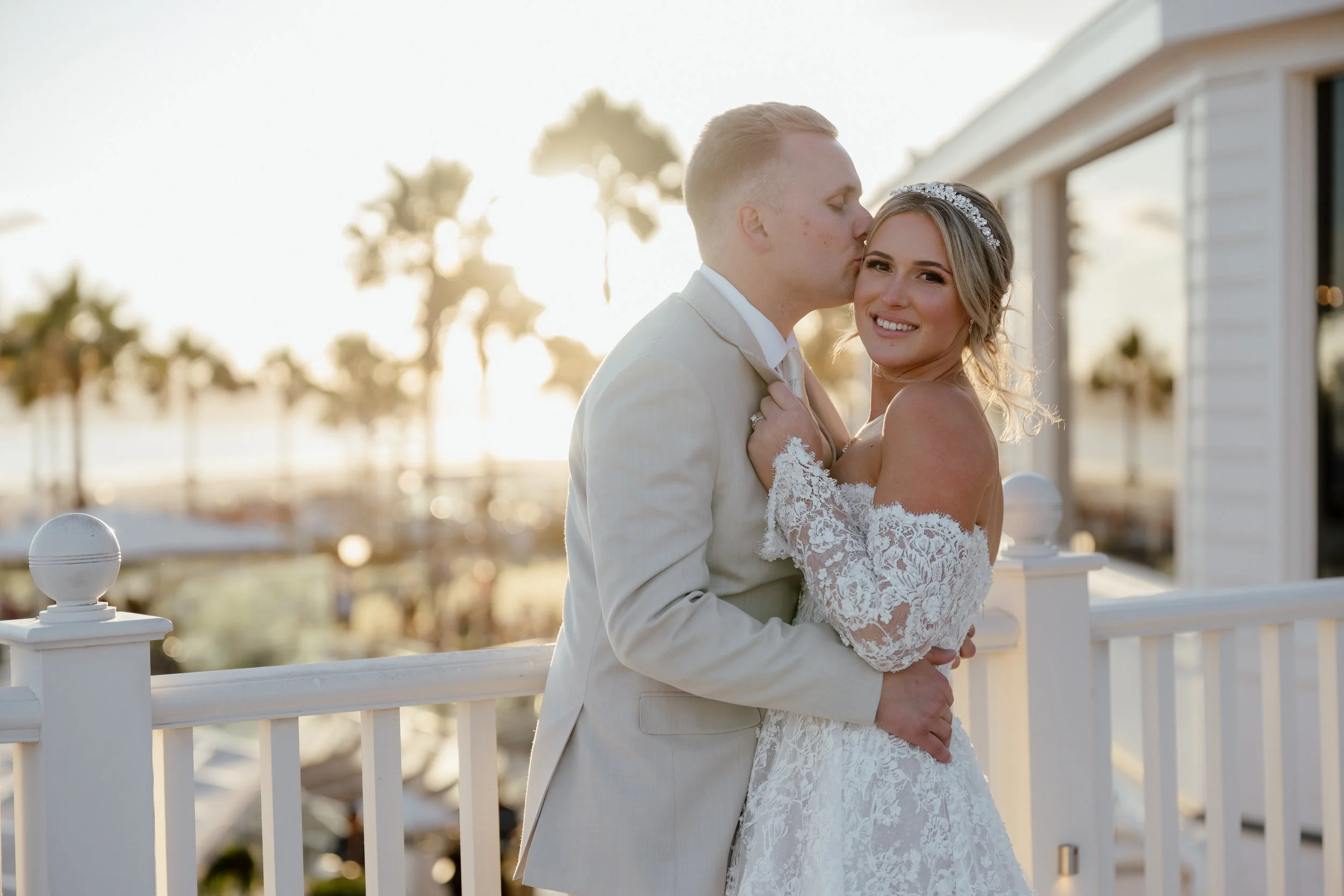 A newlywed couple on a balcony, with the groom kissing the bride's temple and the bride smiling at the camera, during sunset with palm trees in the background at The Hotel Del in San Diego
