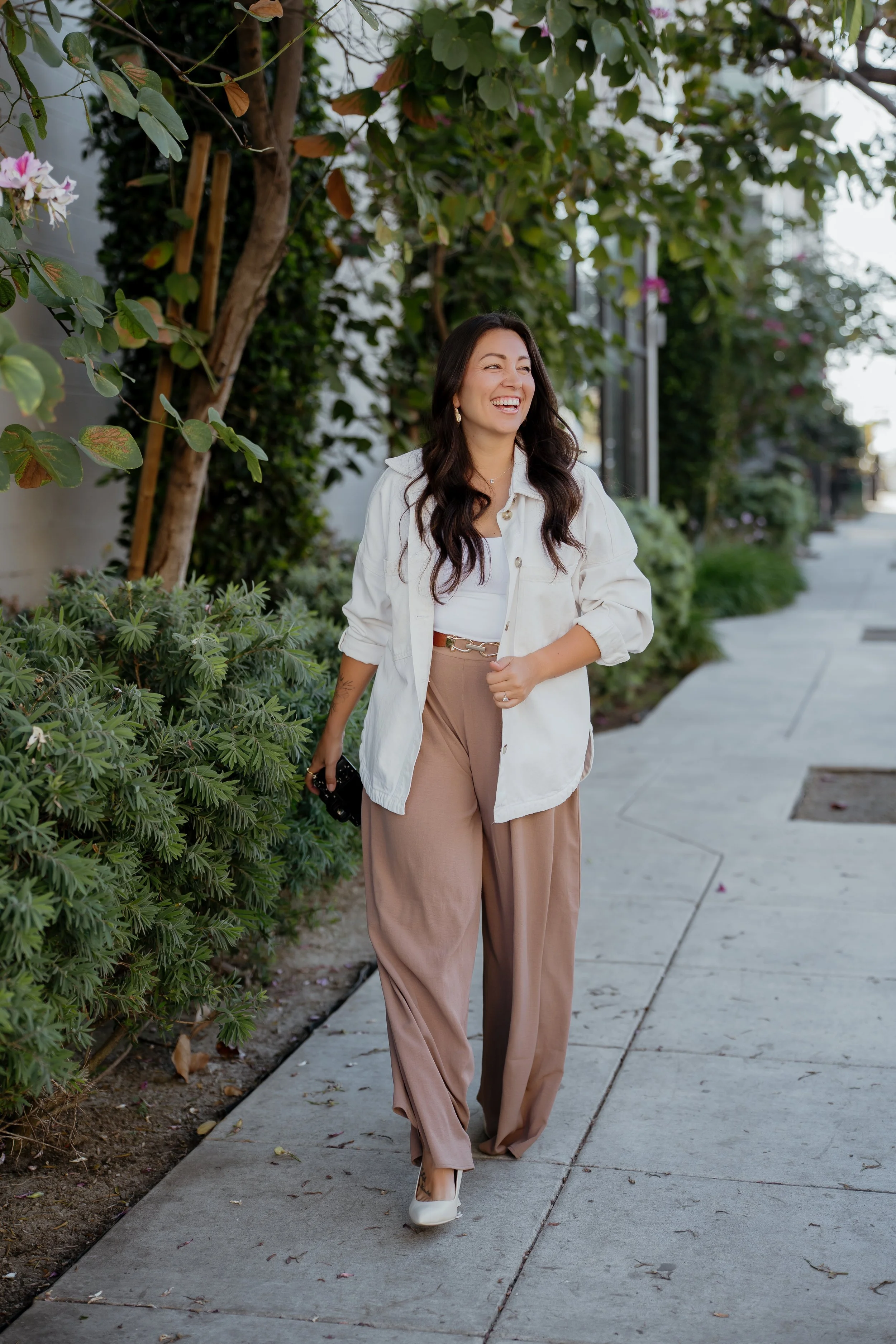 Ally walking on a sidewalk, smiling and wearing a white jacket, beige pants, and white shoes, with greenery and trees in the background.