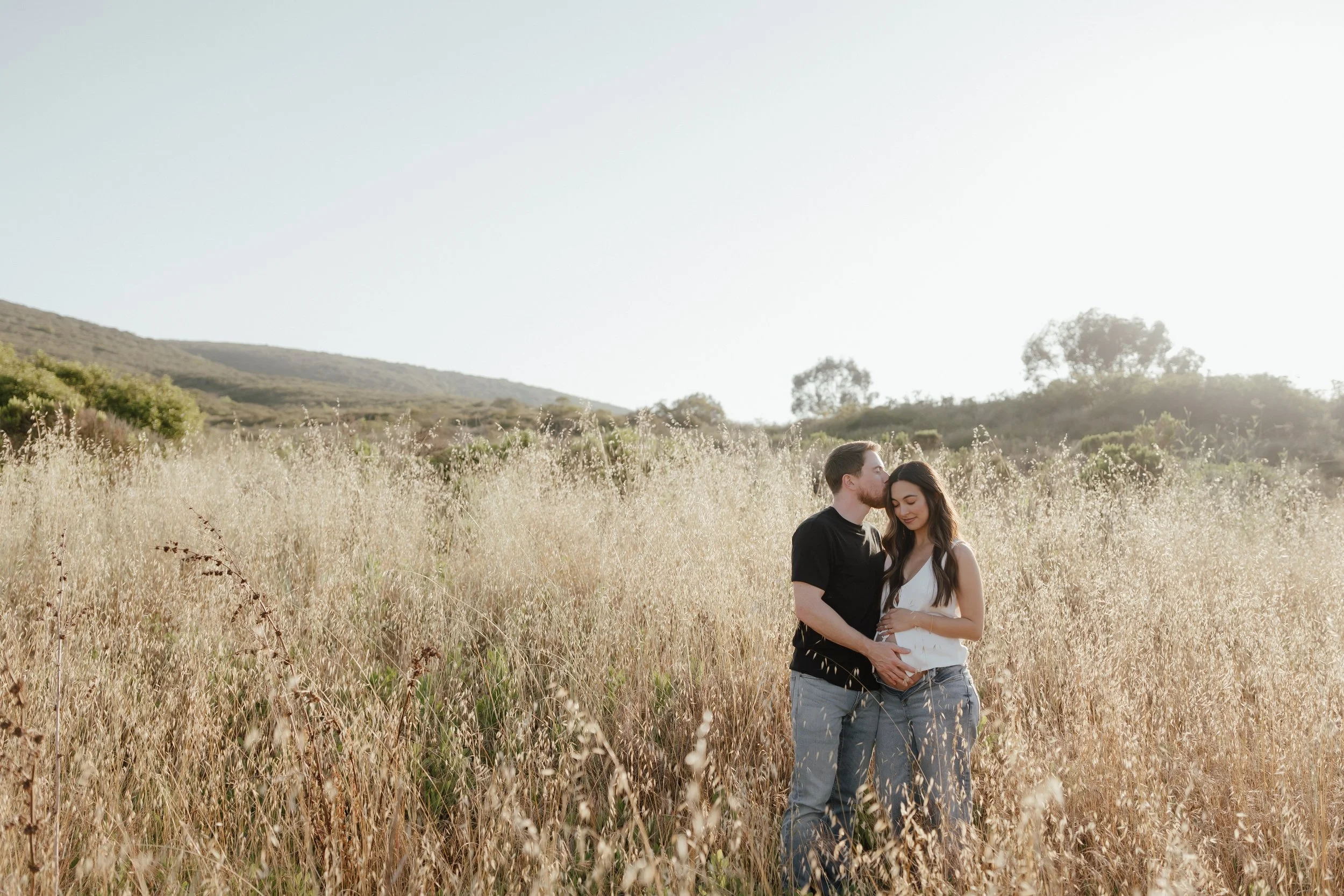 A young couple standing close together in a field of tall grass during sunset or sunrise, with hills and trees in the background.