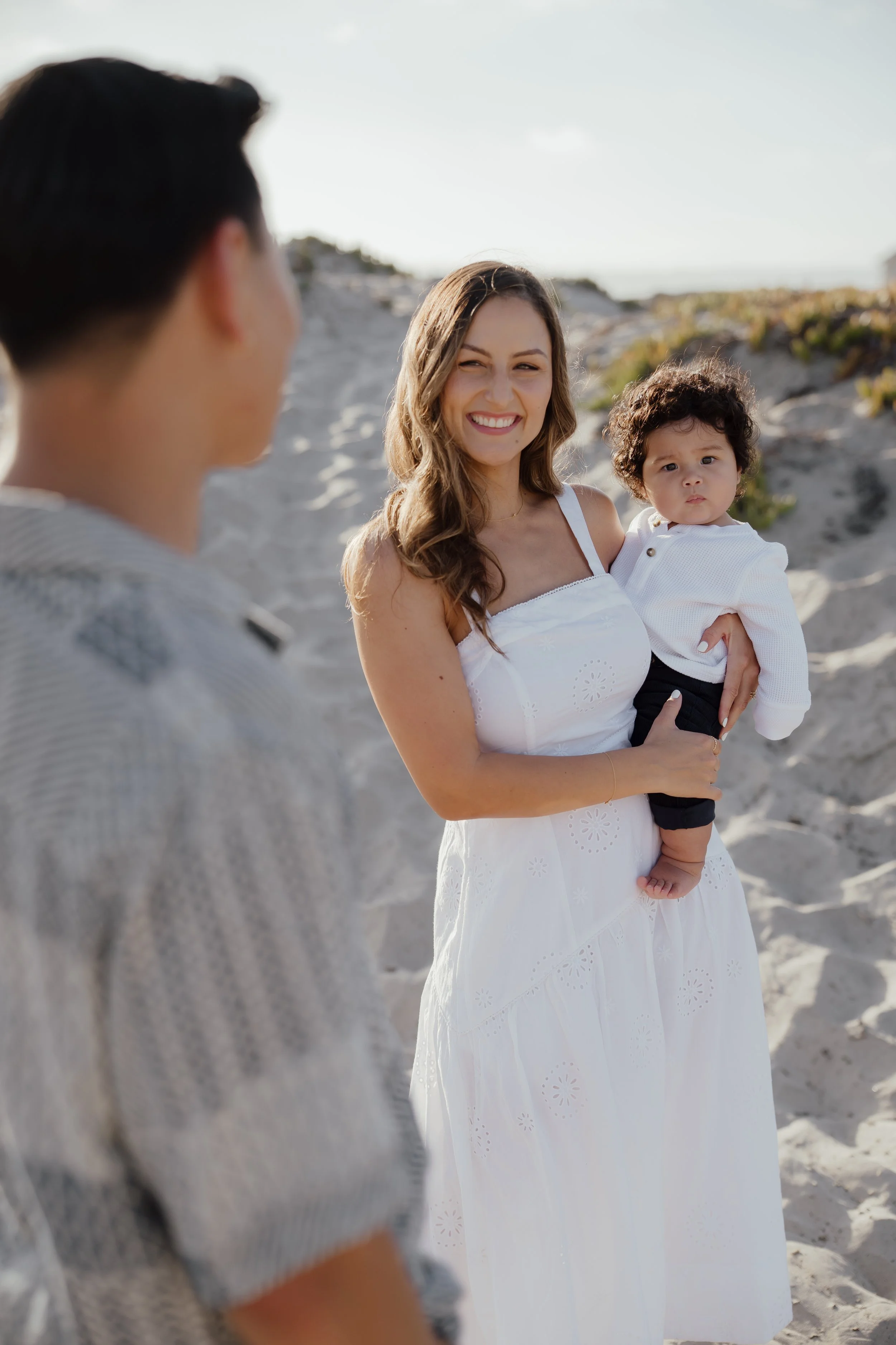 A woman holding a young child on a beach, smiling at a man, with sandy dunes and clear sky in the background.