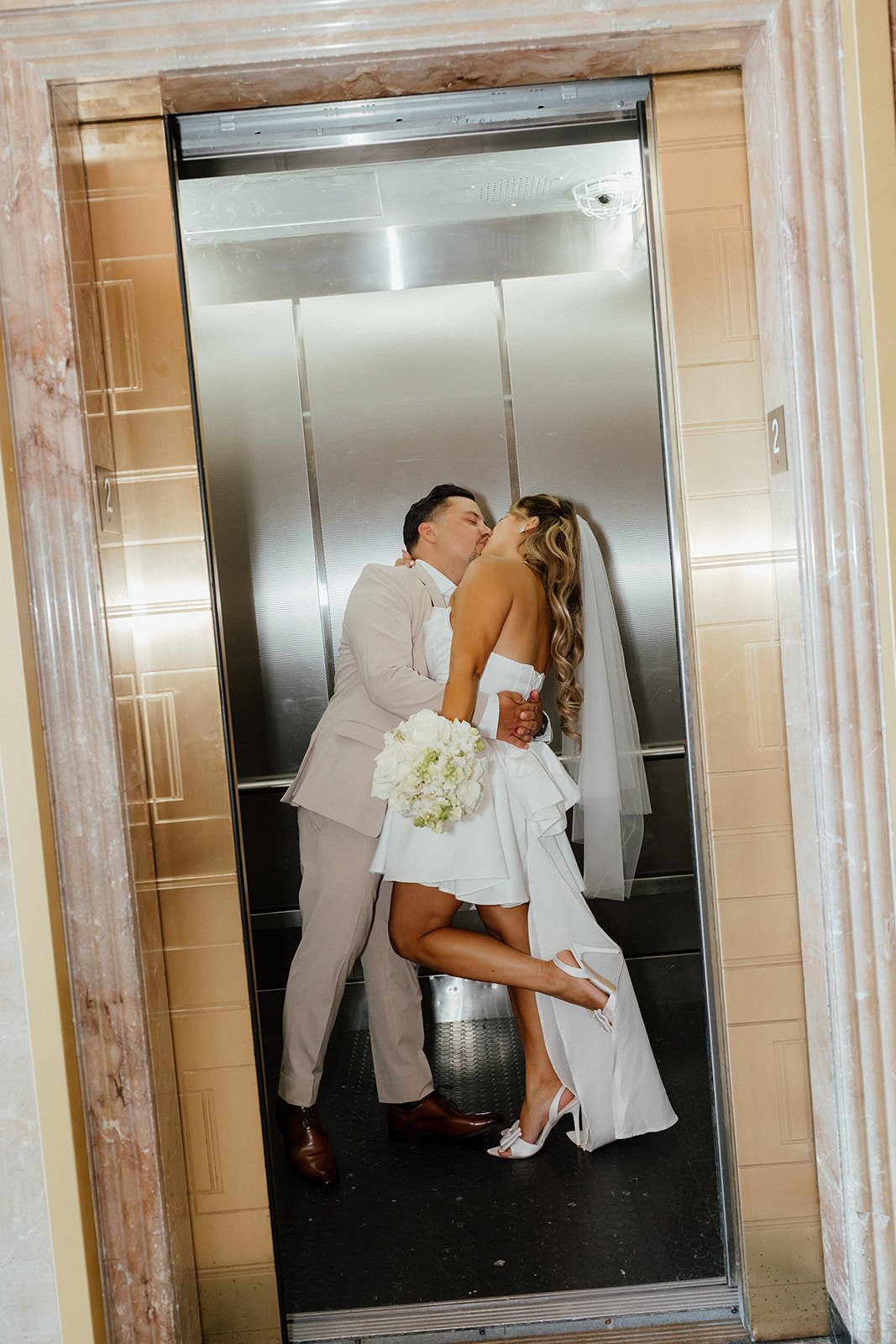 A couple in wedding attire kissing inside an elevator, with the bride holding a bouquet of white flowers during San Diego Elopement.