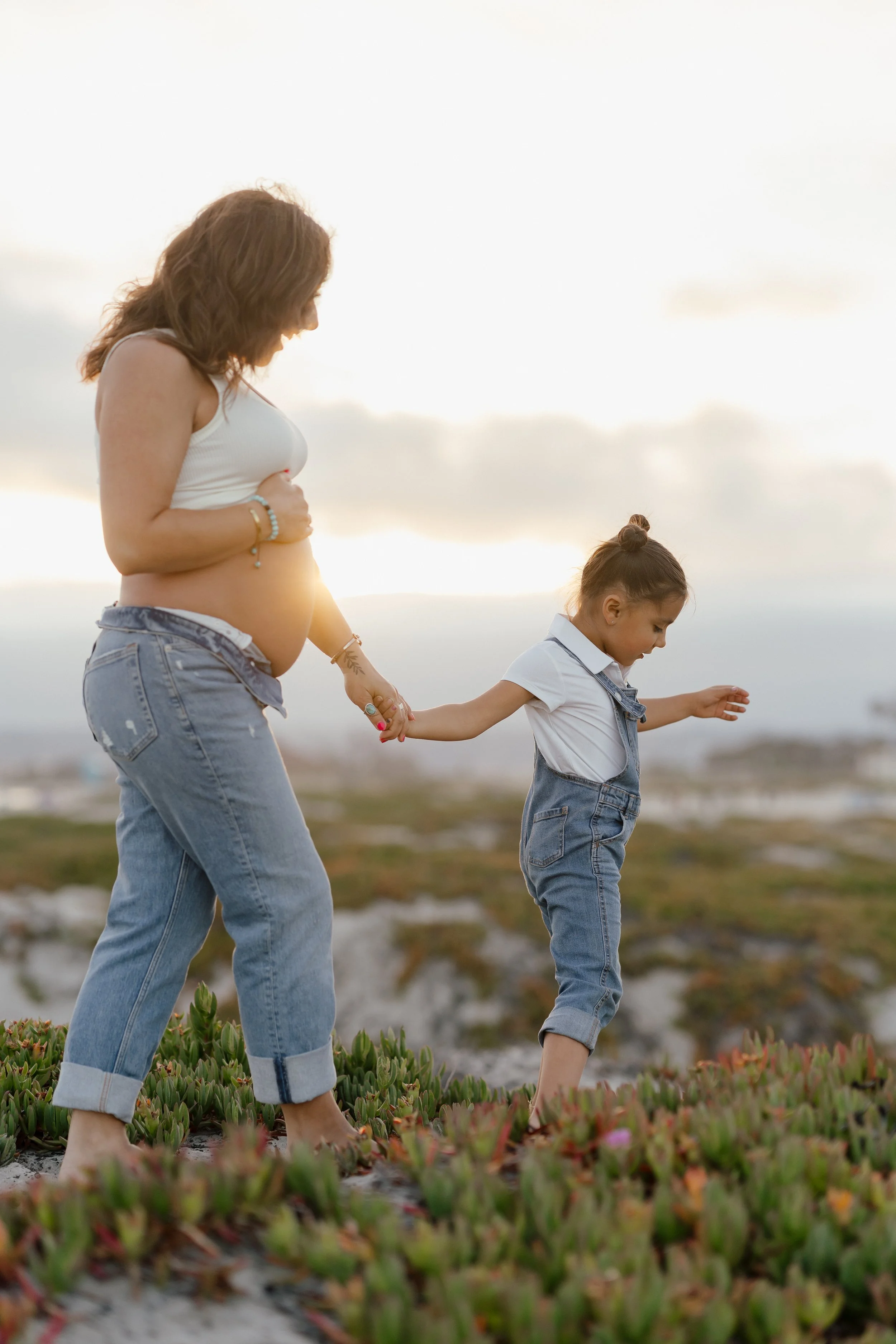 A pregnant woman and a young girl holding hands while standing outdoors on a grassy area during sunset.